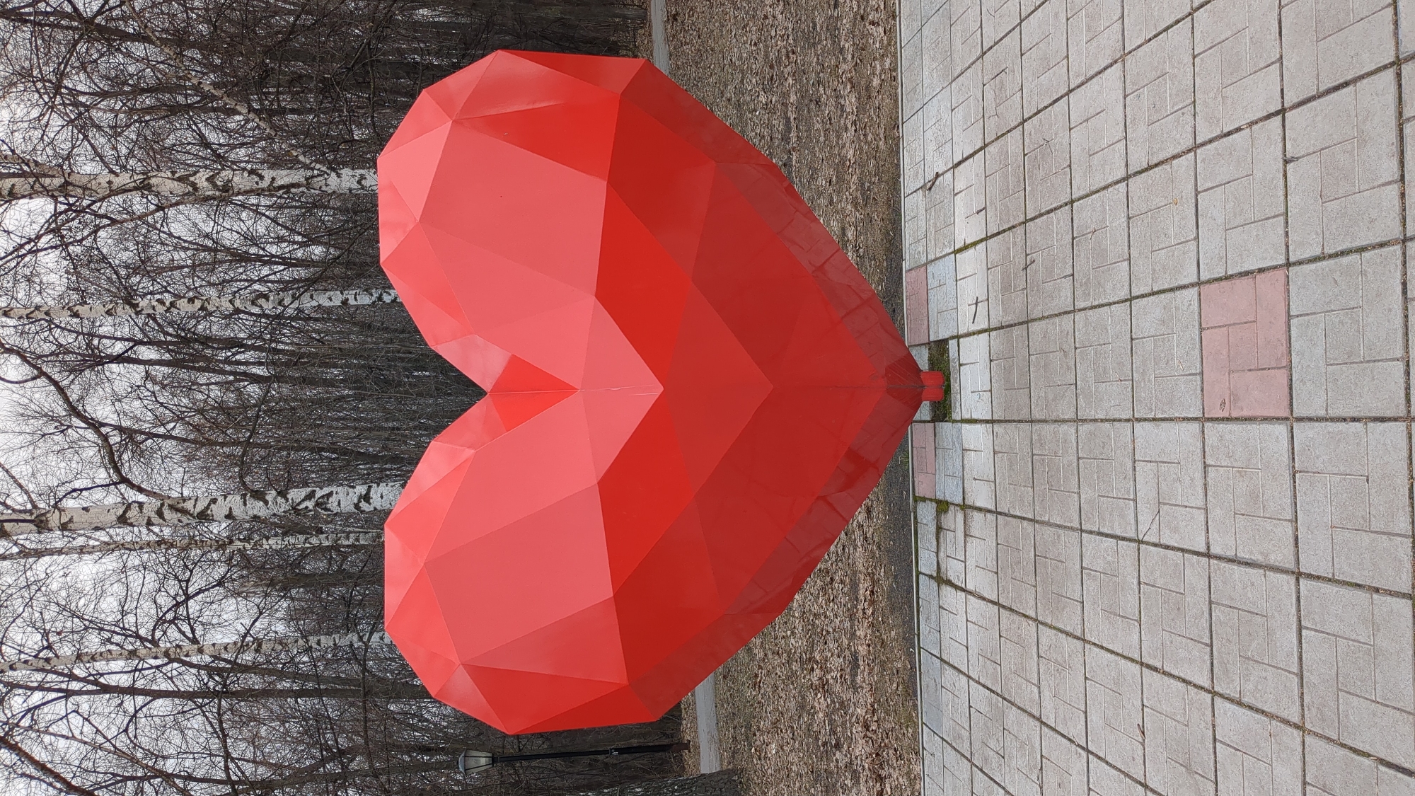A large, red, geometric heart sculpture is situated on a paved walkway in a park in Ulyanovsk, Russia. The heart's base rests on the edge of the walkway, with the other side of the walkway having birch trees in the background. The trees have bare branches, suggesting a late autumn or early spring setting. The walkway is constructed of grey and white rectangular pavers. The photo is taken during the day under overcast conditions.