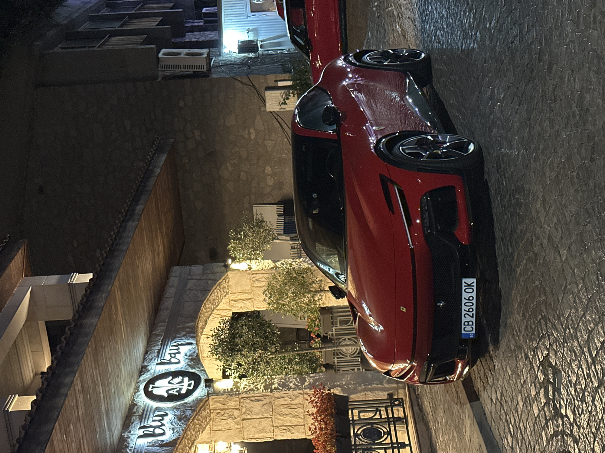 The image depicts a vibrant red Ferrari parked on a cobblestone street at night, directly in front of the "Blu Bay" establishment in Sozopol, Bulgaria. The building facade is constructed of light-colored stone, illuminated by warm lighting, and features a large sign with a circular logo and the words "Blu Bay" in glowing white text. Above the entrance, a wooden awning extends outwards, with visible structural supports.
To the left of the Ferrari, a black metal railing encloses planters filled with red flowers and greenery. Further back, several potted trees with lush foliage are visible near the entrance. The Ferrari itself is the primary subject, showcasing its sleek design, black roof, and prominent front grille. A license plate with the characters "CB 2606 OK" is affixed to the front bumper.
In the background, another red car, partially visible, is parked further down the street. The building to the right of the "Blu Bay" establishment is darker, with visible windows and an air conditioning unit mounted on the exterior wall. The overall scene suggests a sophisticated ambiance, with the luxury vehicle creating a striking contrast against the rustic architecture and evening setting of Sozopol.