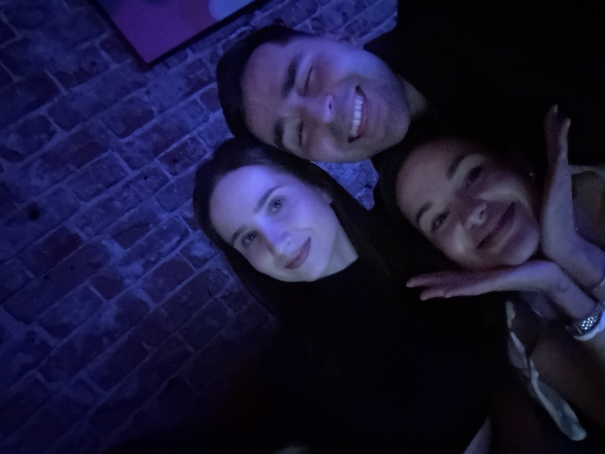 This is a low-light photograph of three people—one man and two women—taken in an indoor setting, possibly a bar or club, in Krasnoyarsk, Russia. The scene is dominated by a strong blue artificial light, suggesting it was taken at night.
The man is positioned at the top right of the frame, smiling broadly with his teeth visible. He has dark hair and is wearing a dark top. To his left, one woman looks towards the camera with a gentle smile; she also has dark hair and is wearing a dark top. The third person, another woman, is at the bottom right, smiling warmly. Her left hand is near her face, and she is wearing a light-colored top and a watch with a metallic strap on her left wrist.
In the background, a textured brick wall is visible, dimly lit by the blue light. All three individuals appear happy and relaxed, suggesting they are enjoying a social gathering. There is no visible text in the image.