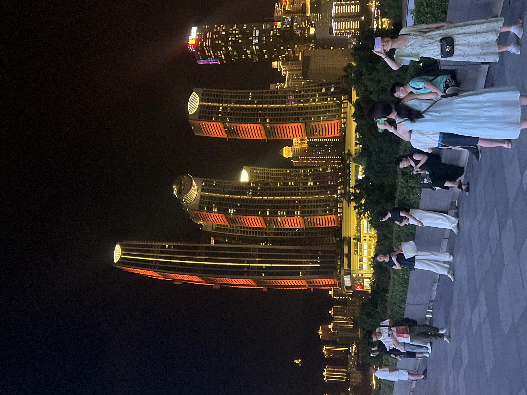 At night, in Jiangbei Cheng Street, China, a group of people stand on a paved walkway facing a city skyline dominated by illuminated skyscrapers. The buildings display vibrant orange and yellow lights, creating a striking contrast against the dark sky. In the foreground, several individuals are casually dressed and engaged in various activities. One woman in a light blue dress holds a phone up, appearing to take a picture or record a video. Another woman in a floral dress is also holding a phone, aiming it towards the skyscrapers. Others are standing or sitting, some looking at their phones. The walkway is bordered by lush green bushes. The scene captures a moment of urban leisure against a backdrop of modern architecture.
