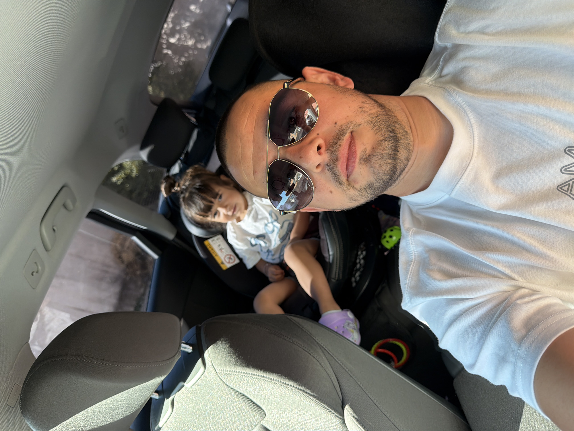 A man and a young child are in the back seat of a car. The man is in the foreground, looking at the camera and wearing sunglasses and a white t-shirt. The child is in a car seat behind him, wearing a white t-shirt and purple shoes. The car appears to be parked on a street in Burgas, Bulgaria, based on the visible foliage and building styles. The scene suggests a family outing or trip.