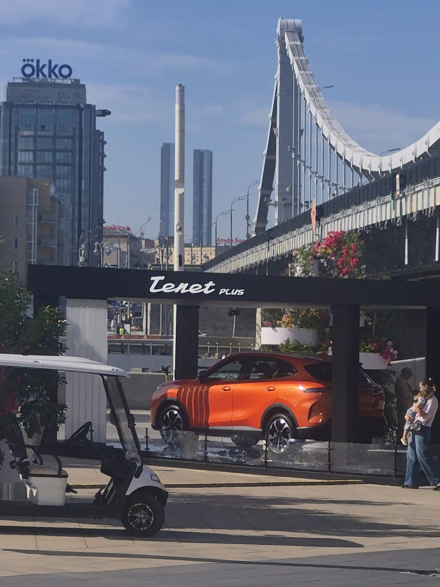 In an urban environment under a clear blue sky, a bright orange SUV is prominently displayed on a raised platform. The vehicle is sleek and modern, with a distinctive body design and eye-catching alloy wheels. Above the car, a black canopy features white lettering that reads "Tenet PLUS," suggesting a brand or promotional event. To the left of the SUV, a white golf cart with a black roof and canopy is parked, its wheels visible.
In the background, a large suspension bridge dominates the skyline, its complex structure of cables and towers a testament to engineering. Tall modern buildings, including one with the illuminated blue logo "ökko," rise behind the bridge, indicating a bustling city center. The scene is likely set outdoors, possibly a plaza or public space, given the presence of a pedestrian walkway in the foreground.
A woman, holding a child, walks to the right of the frame, partially obscured by a dark vertical support structure. Her blue jeans and light-colored top suggest casual attire. Another person, a man, is visible in the far right background, appearing to be walking away from the camera. The overall atmosphere is one of a bright, sunny day in a modern city, with a focus on automotive display and urban architecture.