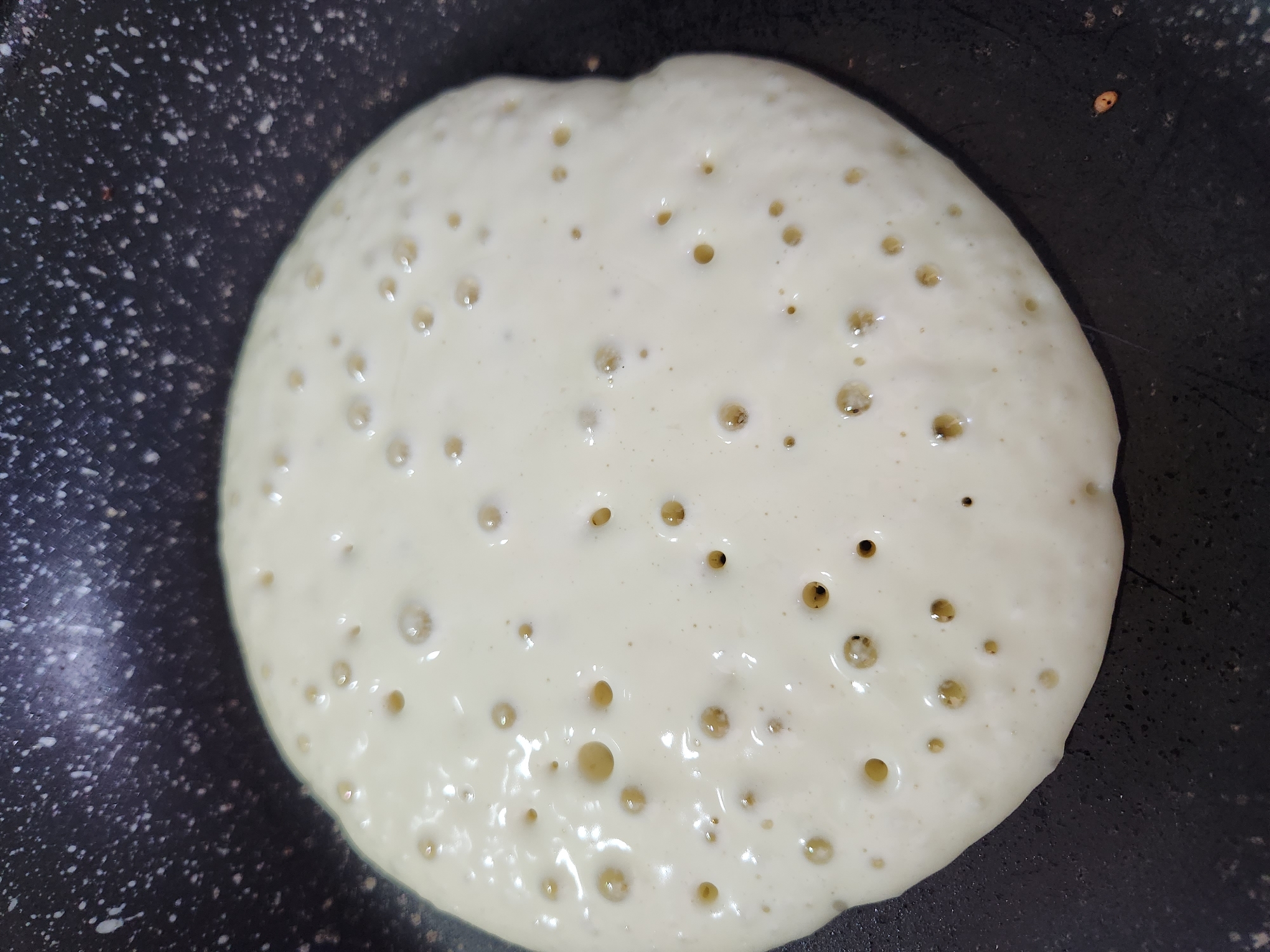 A close-up, top-down view of a pancake being cooked in a non-stick frying pan. The pancake is light yellow, with a slightly irregular circular shape. Bubbles are visible on the surface of the batter, indicating it is cooking. The frying pan is black with a speckled pattern and has a slightly worn appearance. The lighting suggests an indoor setting. The image does not contain any elements that would place it in a specific city like Đà Nẵng, Vietnam.