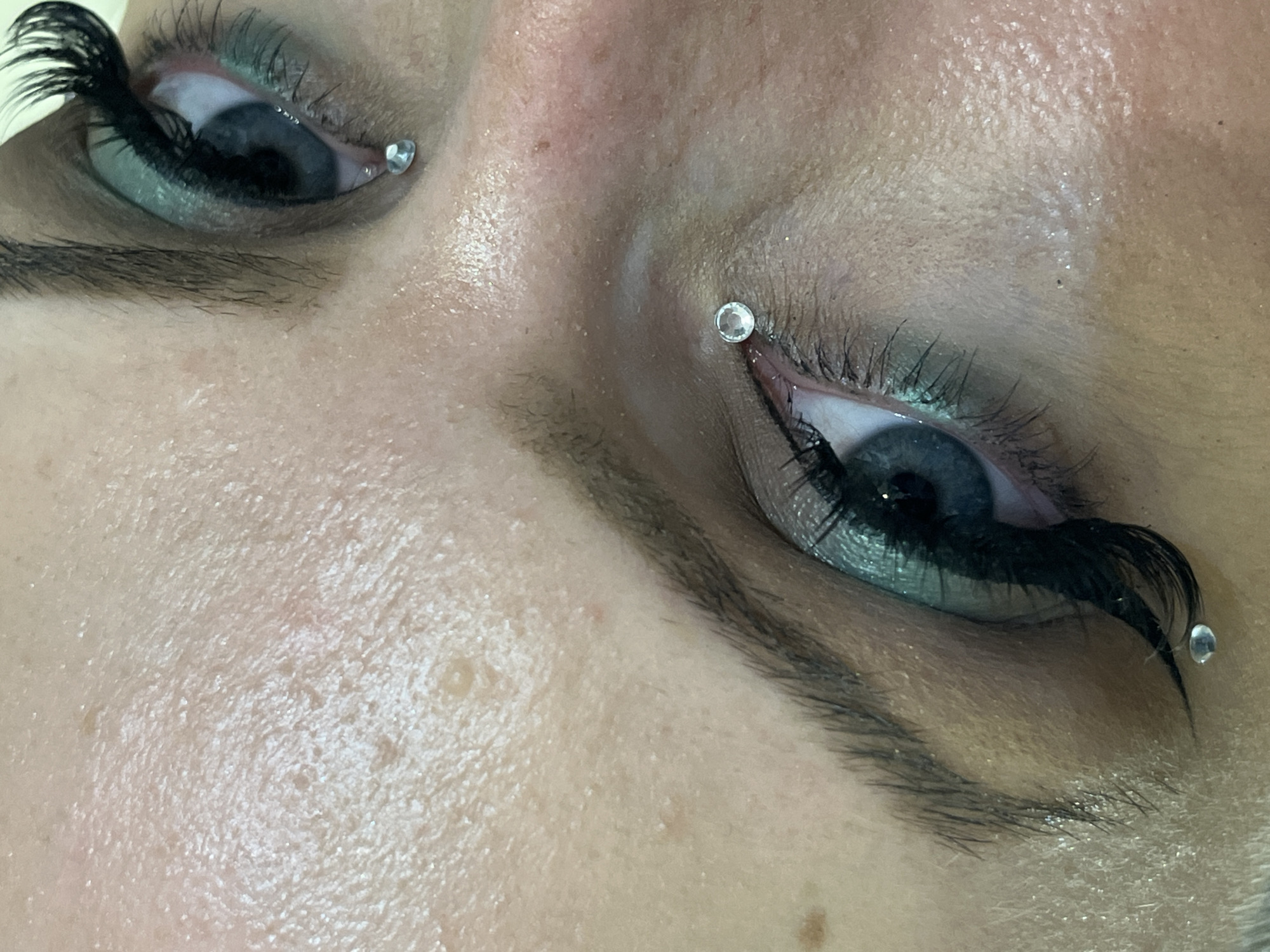 The image depicts a close-up of a person's eyes with elaborate makeup. The eyes are blue, adorned with false eyelashes and what appears to be light green eye shadow. Small, round, clear gemstones are attached near the corners of both eyes. The skin around the eyes is fair with visible pores and slight imperfections. The eyebrows are dark and thick.
There is no information available in the image to associate this scene with the city of Plovdiv, Bulgaria.