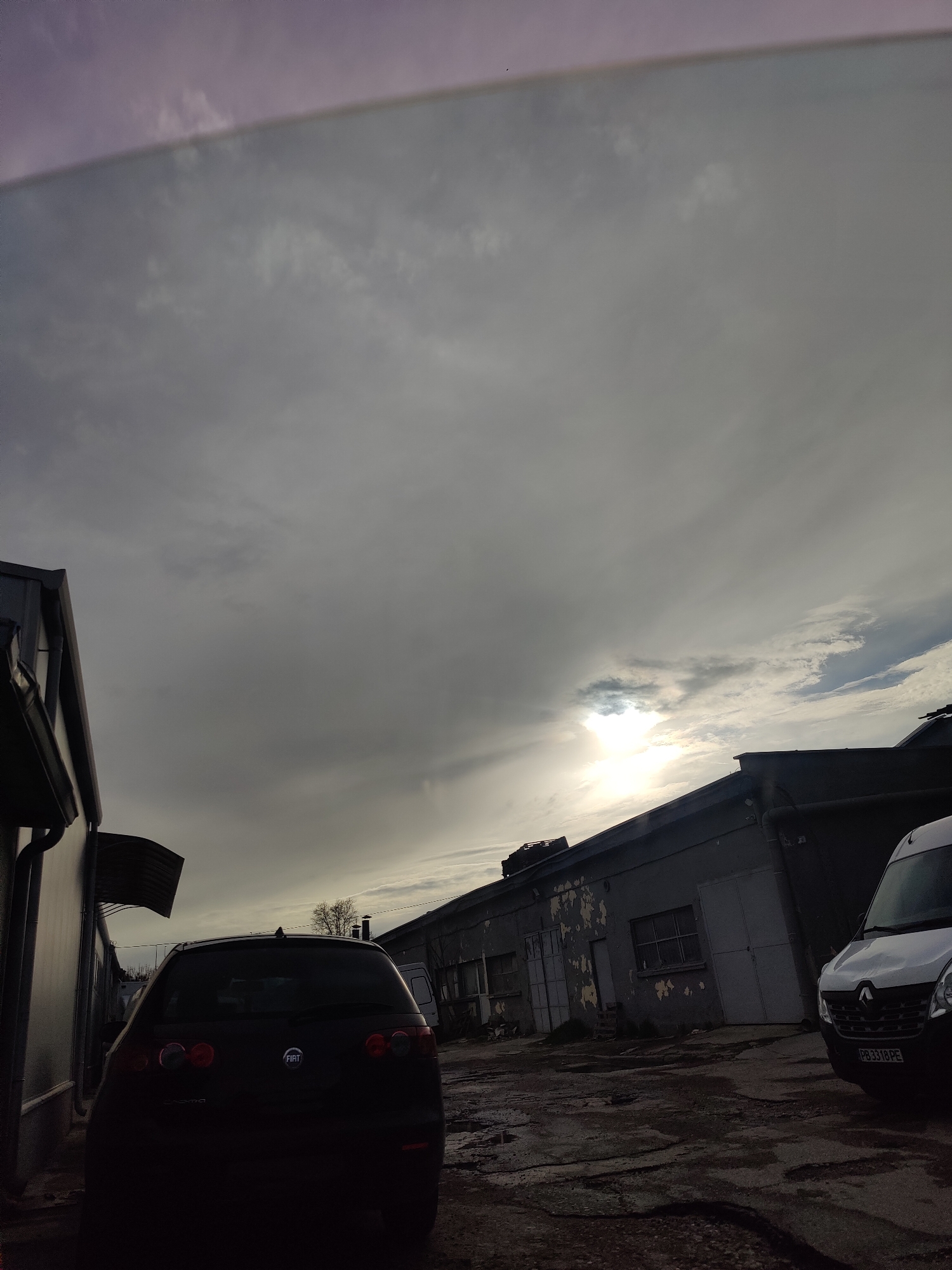 The photograph appears to be taken from within a vehicle, looking out onto an industrial or commercial area in Plovdiv, Bulgaria. The sky is overcast, with the sun partially obscured by clouds.
In the foreground is a dark-colored Fiat Croma hatchback, parked next to a building with a rain gutter. To the right of the Fiat, there is a white Renault van with the license plate "PB 3318 PE".
The buildings in the background are single-story industrial structures with peeling paint and closed metal doors. The ground is paved, with visible cracks and patches. In the distance, a bare tree is visible.