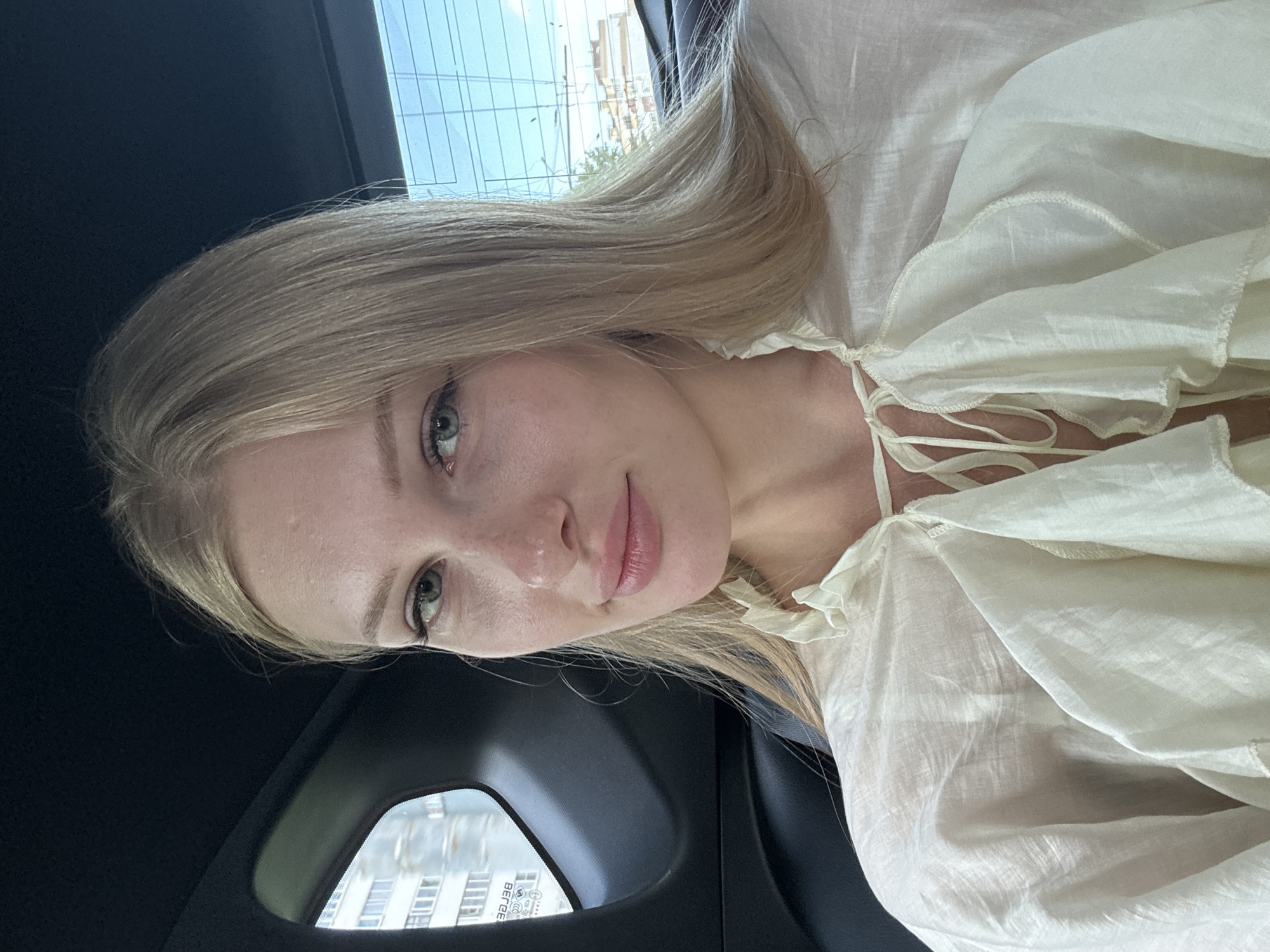 A young woman with blonde hair and green eyes is taking a selfie in the back seat of a car. She is wearing a light-colored, ruffled blouse. The car's interior, including black leather seats and a dark headliner, is visible. Through the windows, a building and a blue sky with some trees are seen, suggesting an urban setting. The background details, such as the architecture visible through the windows, are consistent with an area within the city of Kazan, Russia.