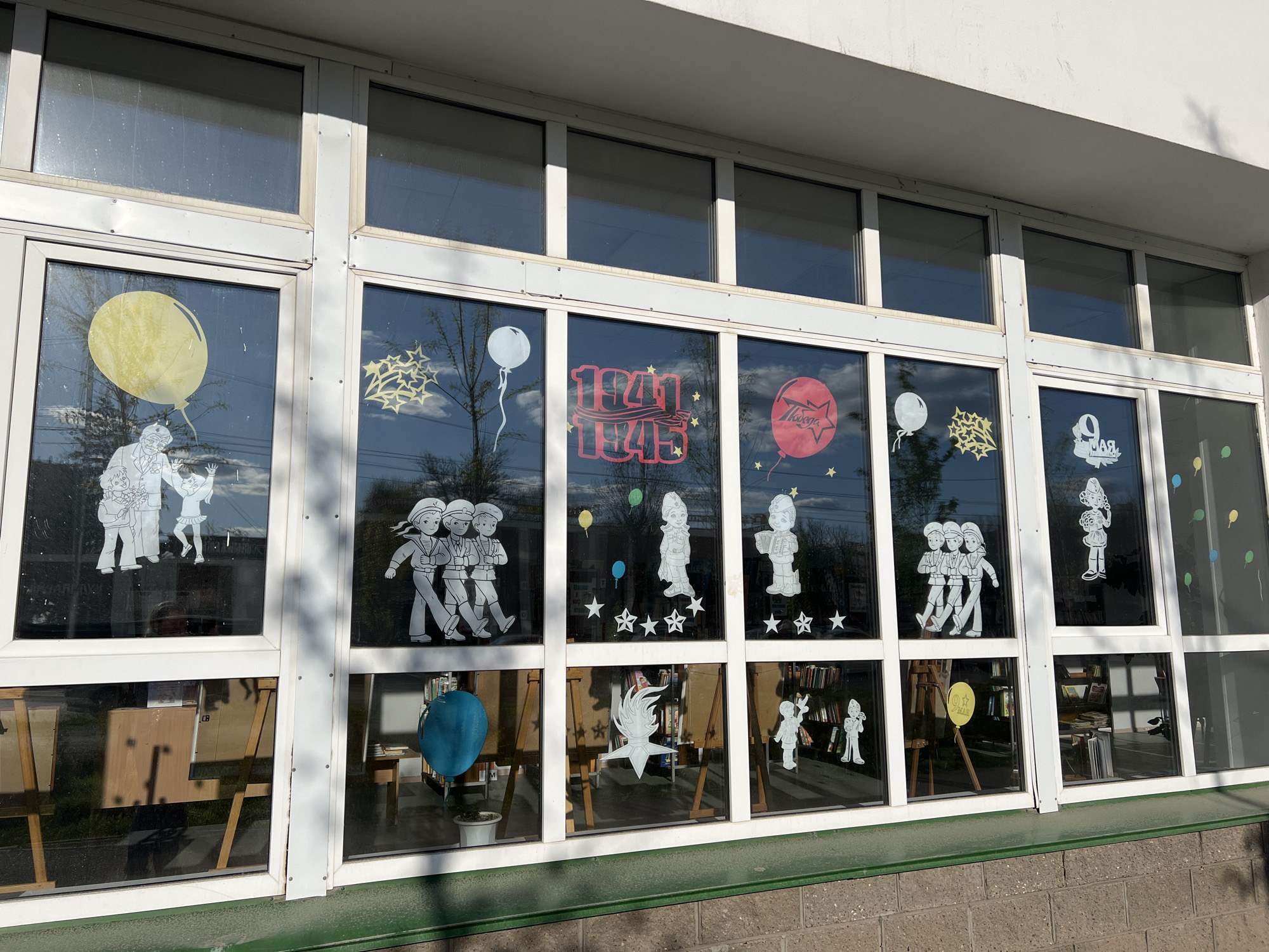 A view of a building's large windows decorated with white paper cutouts, likely in Ufa, Russia. The cutouts commemorate a historical event, possibly Victory Day (May 9th). They depict soldiers, happy children, balloons, stars, and the years "1941-1945". The inside of the building appears to be a library or classroom. Some easels and bookshelves are visible. The window frames are white. The outside ledge below the windows is painted green. The lower portion of the building’s wall is made of gray bricks. The sky reflecting in the windows suggests it is daytime.
