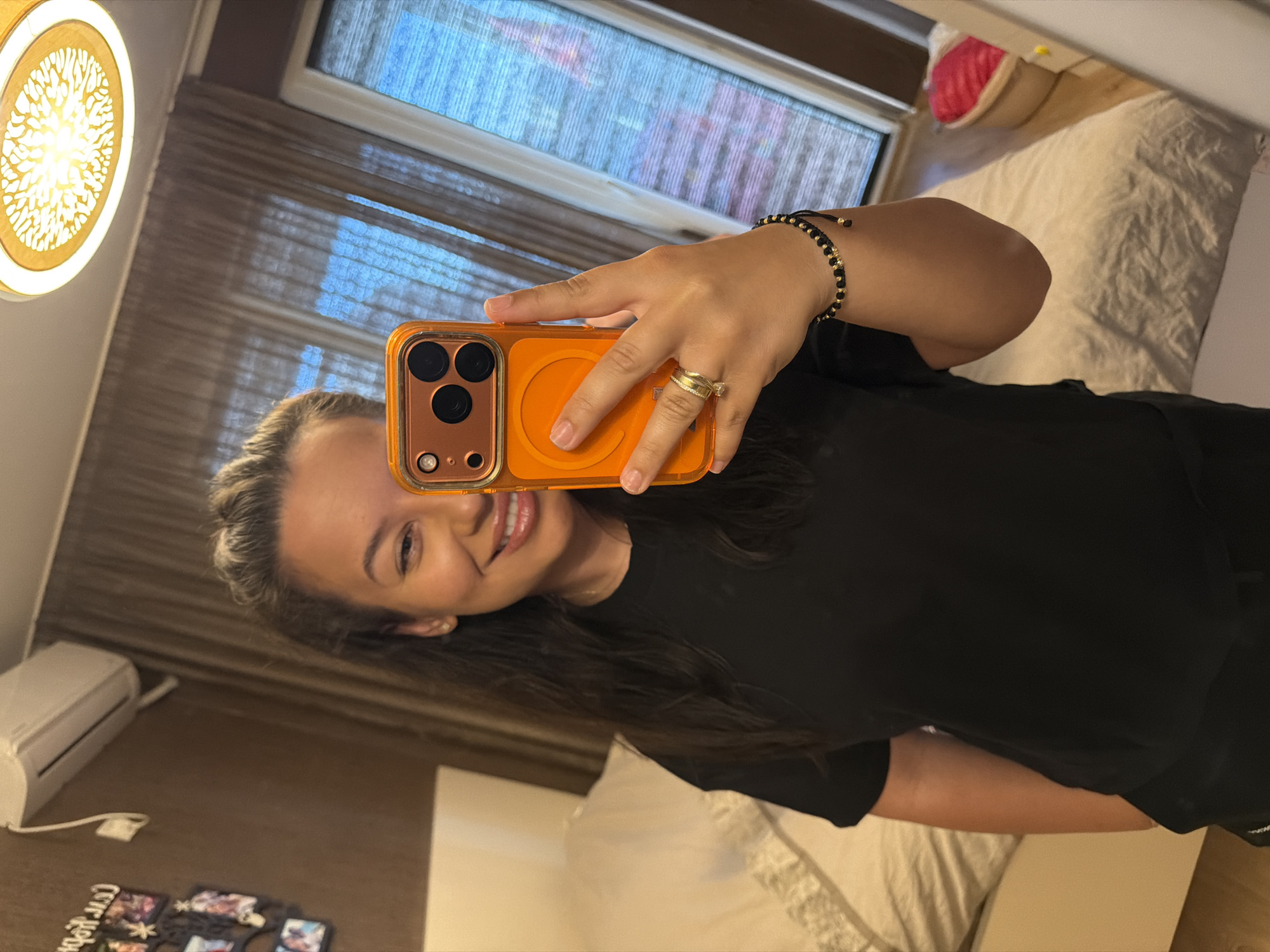 A woman is taking a selfie in a bedroom. She is holding an orange phone in her right hand, which is positioned in front of her face, obscuring the left side of her face. She is smiling and looking into the phone's camera. She has long, dark hair that is pulled back from her face. She is wearing a black t-shirt and a ring on her left ring finger. A bracelet with black and gold beads is on her left wrist. The room appears to be a bedroom with a bed in the background. The walls are a warm brown color, and there are curtains on a window. An air conditioning unit is mounted on the wall. A decorative frame with multiple photos is on the left wall. The lighting in the room appears to be a mix of artificial and natural light. The overall setting suggests a casual, personal environment. There is no visible indication of the location being in Burgas, Bulgaria.