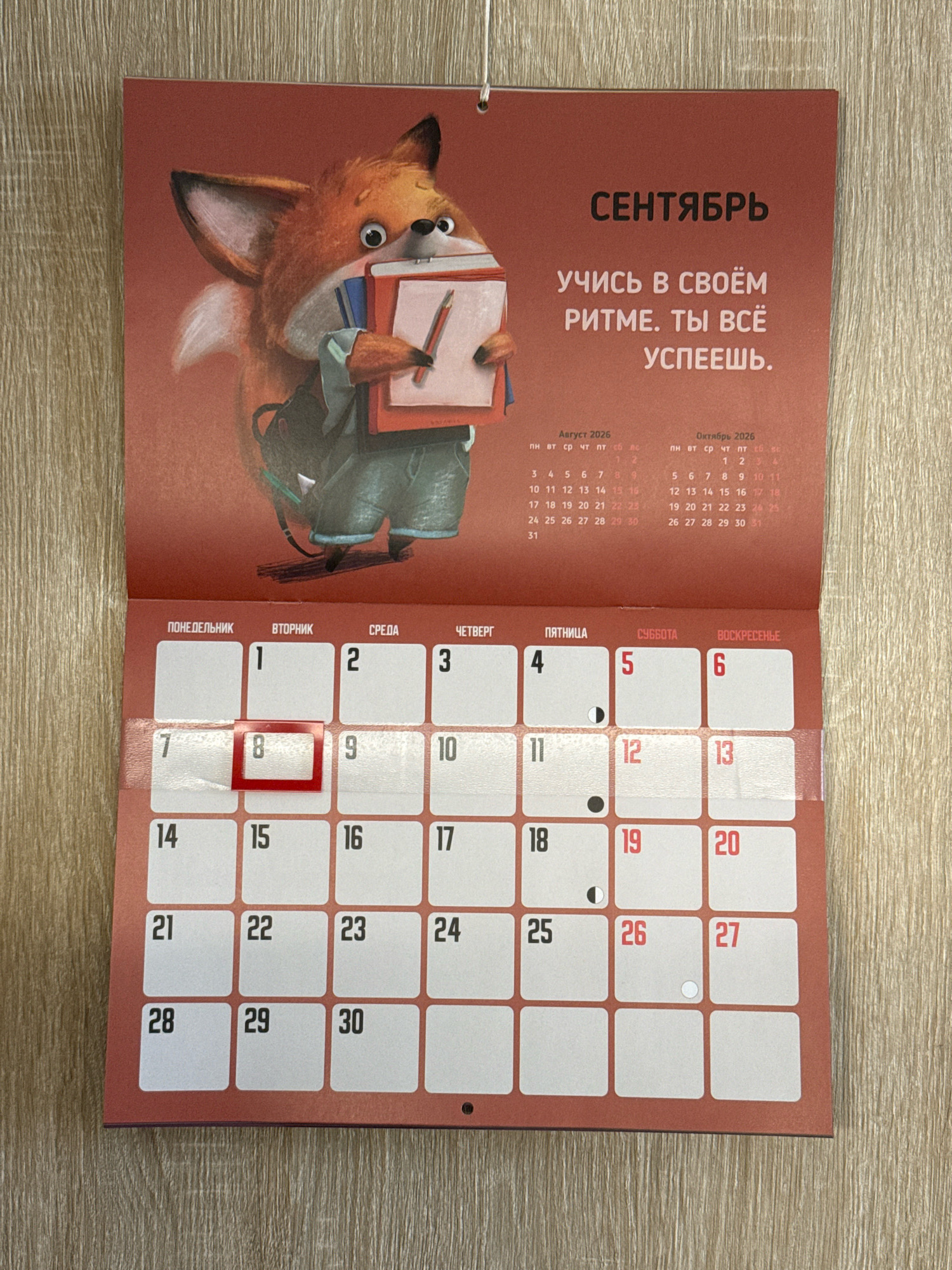 The image shows an open calendar with a large illustration of a cartoon fox holding books and a notepad. The calendar is for September and the text above the fox says "September" in Russian. The calendar grid below is for the month of September, with days of the week across the top: "Monday", "Tuesday", "Wednesday", "Thursday", "Friday", "Saturday", "Sunday". The dates are visible in the grid. The calendar is positioned on a wooden surface. There is no visible indication of the city of Lyubertsy, Russia.