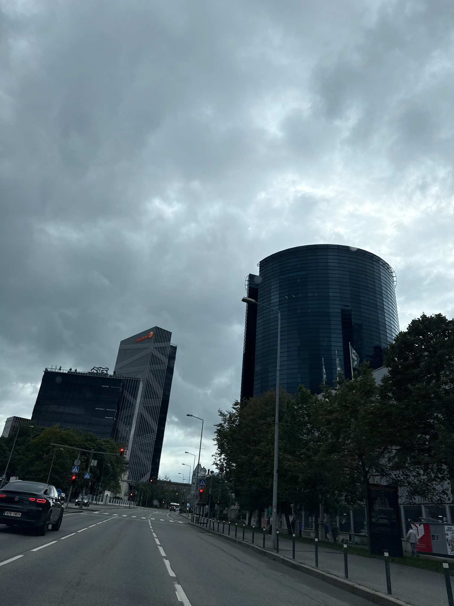 The image captures a street view in Tallinn, Estonia, on an overcast day. Dominating the skyline are modern architectural structures. To the right, a tall, cylindrical building with a dark, reflective facade is visible. To the left, two darker, angular skyscrapers stand side-by-side; the left one has "ATR" signage, and the right one displays an orange logo. A black car with a license plate reading "676 H-KH" is driving on the asphalt road, which has clear lane markings and traffic lights. Several trees line the street, with some foliage visible on the buildings themselves. Pedestrians are sparsely scattered along the sidewalks. The sky is filled with heavy, dark clouds, suggesting the possibility of rain.