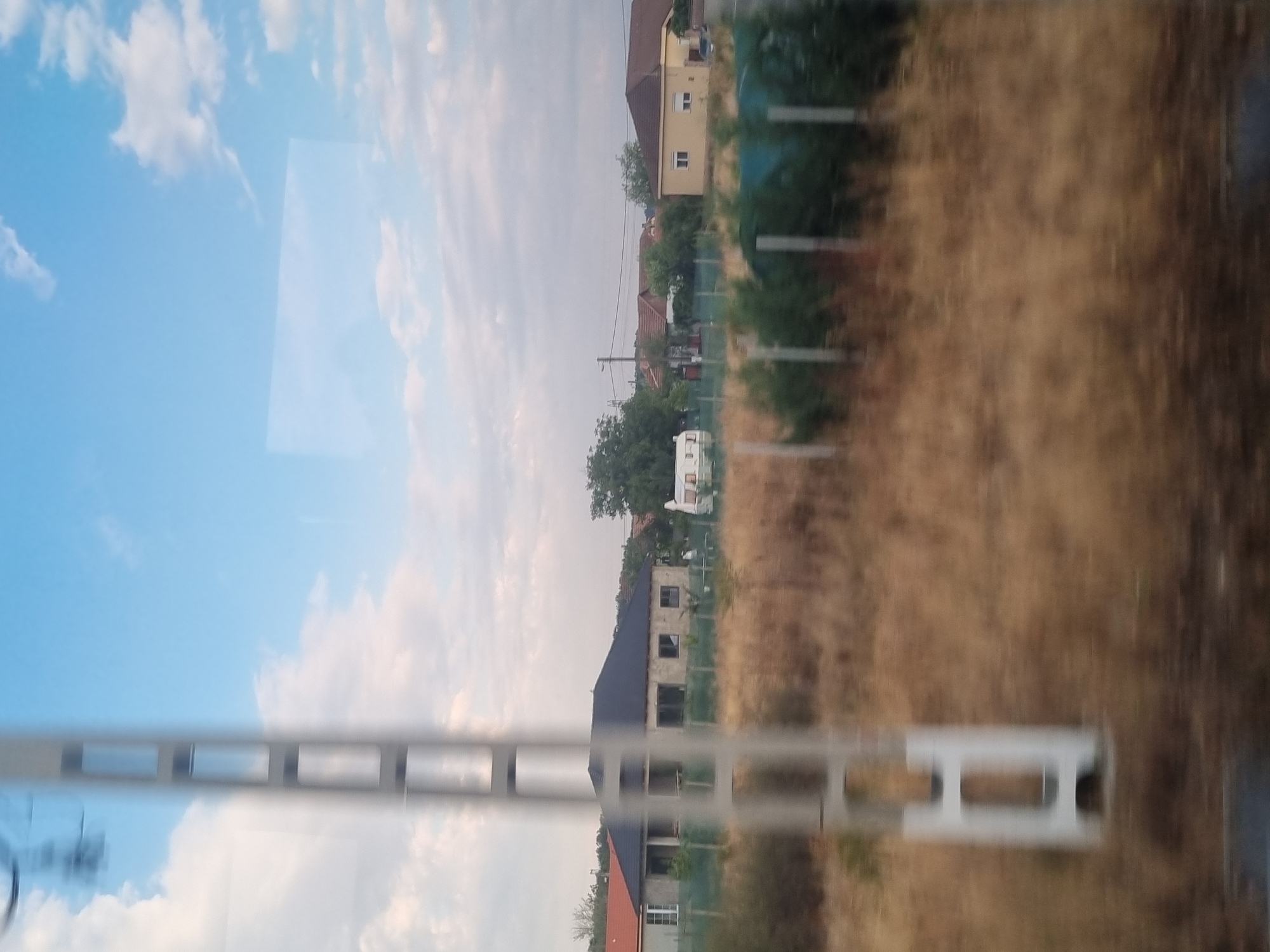The image shows a blurry, outdoor scene with a focus on residential buildings and a large utility pole in the foreground. The sky is blue with scattered white clouds. In the middle ground, several houses with tile roofs are visible, along with some trees and a white trailer or caravan. The ground is covered in dry, brown grass. The scene appears to be taken from a moving vehicle, as indicated by the motion blur. The location is identified as Gepiu, Romania.