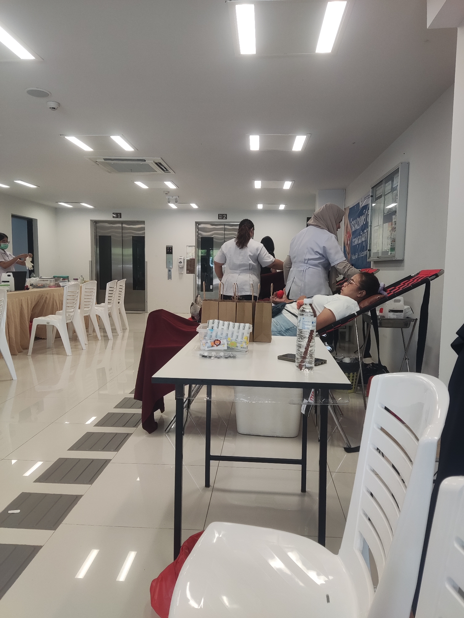 The image shows a room, likely in a health center or clinic in Baan Bo Phut, Thailand, during a blood donation event. Several people are visible, including at least two healthcare workers in white coats and a woman lying on a reclining chair, presumably donating blood. The room is brightly lit by fluorescent lights. Two elevators are visible in the background, labeled with the number "1" and "2". Tables are set up with supplies like water bottles and packaged items. White plastic chairs are scattered around the room. The floor is tiled with a patterned design that appears to be raised tactile paving. A bulletin board with documents is affixed to the wall.