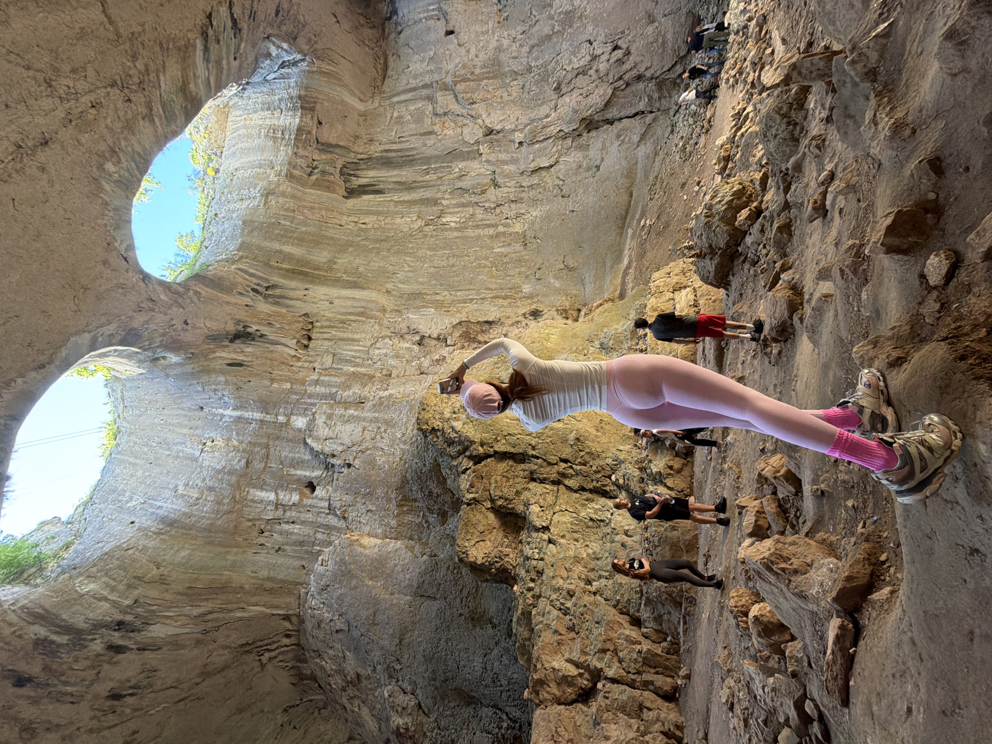 The image shows the Prohodna cave near Karlukovo, Bulgaria. The cave's interior is lit by natural light entering through two large openings in the ceiling. A group of people are standing on the cave floor, with a woman in pink leggings and a white shirt closest to the camera, apparently taking a selfie. Other individuals are nearby. The cave walls are textured and rocky.