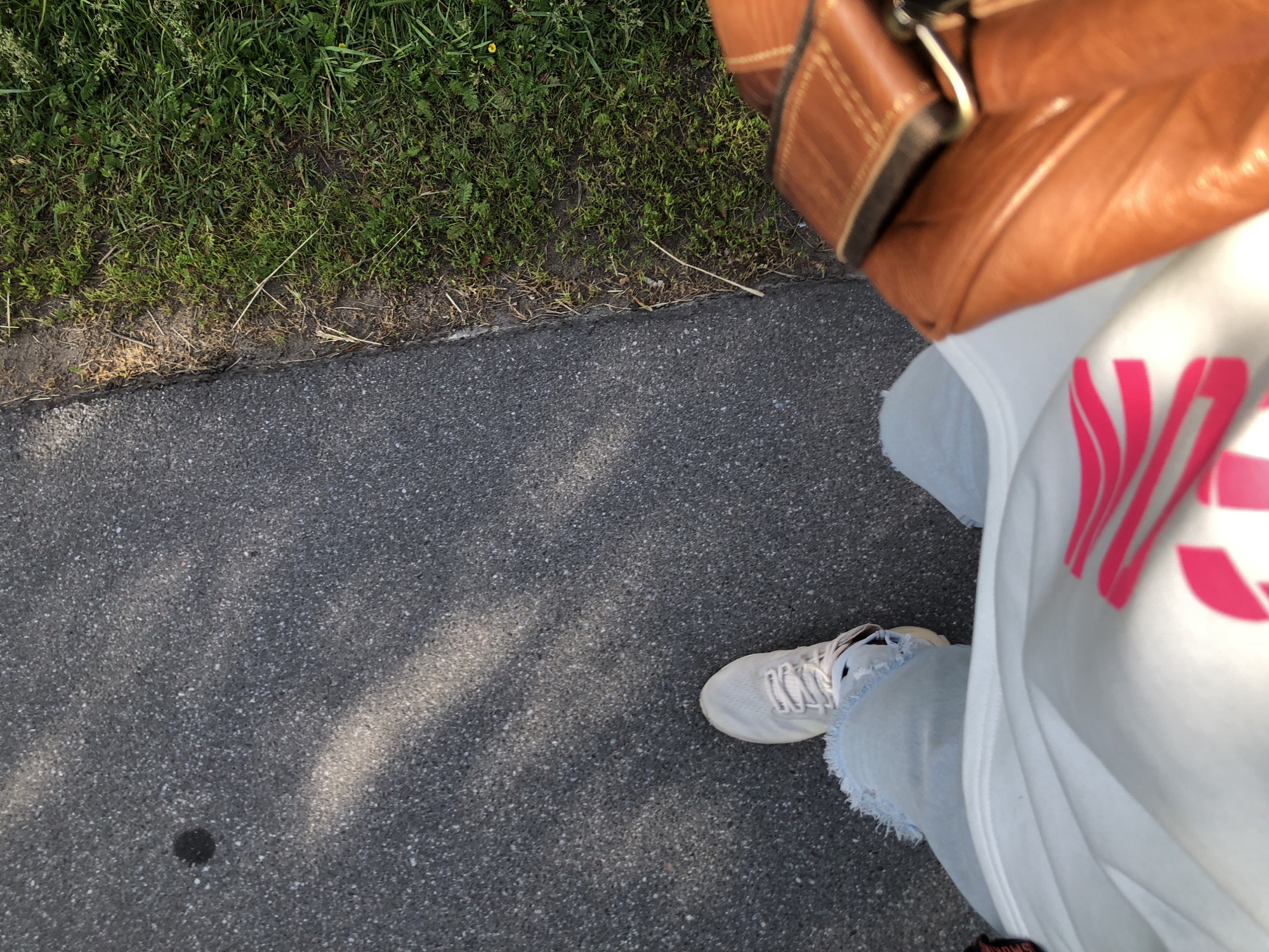 The image shows a first-person perspective of a person walking on a paved path next to a grassy area. The person is wearing light-colored sneakers, ripped light-blue jeans, and a white sweatshirt with a bright pink graphic design. A tan leather bag is visible on the right side of the frame. The path appears to be asphalt with some shadows cast from an unseen source. The grassy area is green and somewhat uneven, with some dry grass and small yellow flowers. The context of Saint Petersburg, Russia, is not visually evident in the image itself, as it lacks any identifiable landmarks or signage specific to that location.