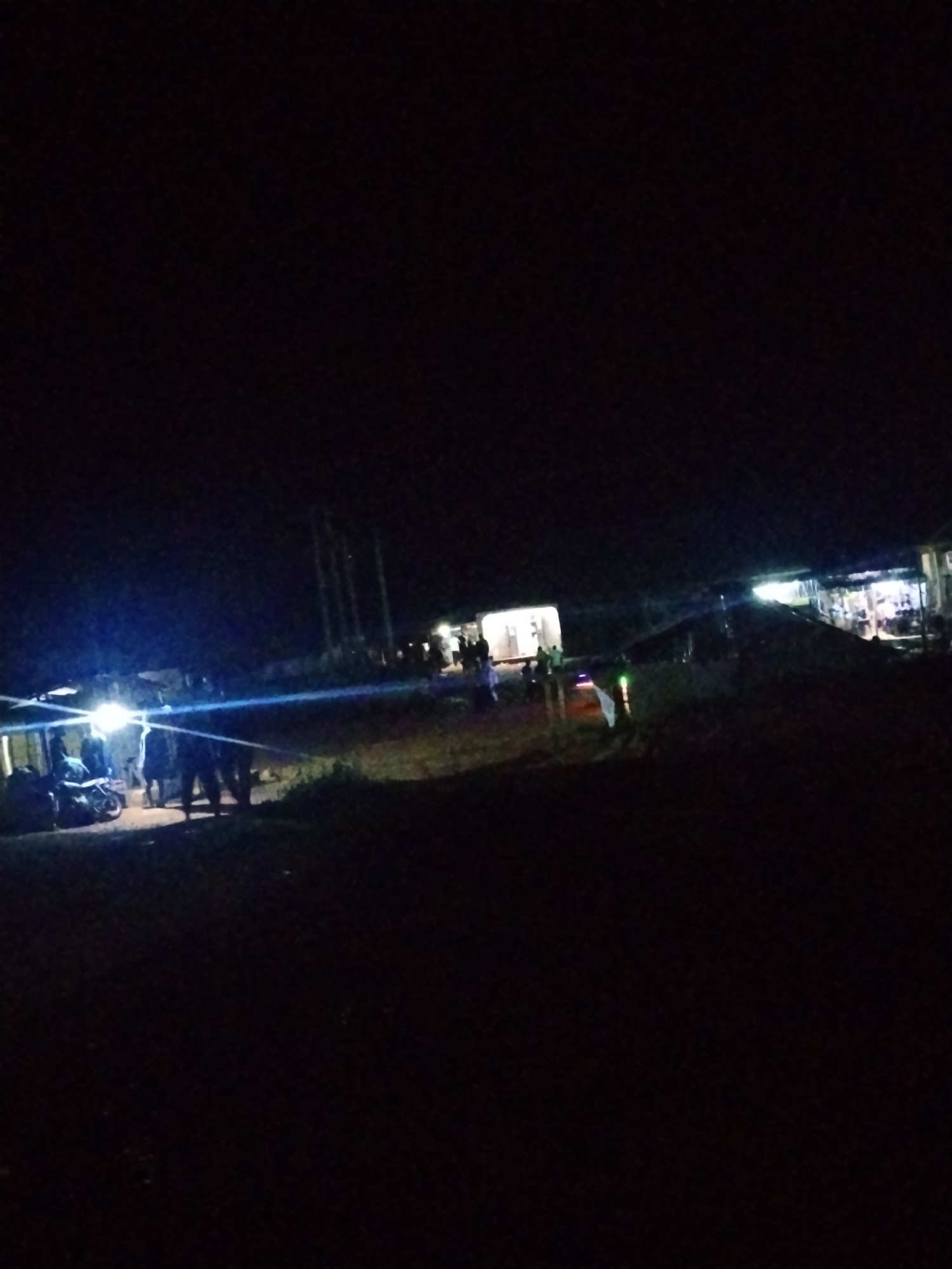 The image depicts a nighttime scene in Durumi, Nigeria, likely a semi-urban or rural area. The foreground is dark, with some uneven terrain visible. In the mid-ground, a bright light source, possibly from a vehicle's headlight, illuminates a group of individuals standing near what appears to be a motorcycle. Further in the background, several structures with lights are visible, suggesting buildings or makeshift shops. A few more people are scattered around these illuminated areas. The overall impression is of a gathering or activity taking place after dark, illuminated by artificial light.
