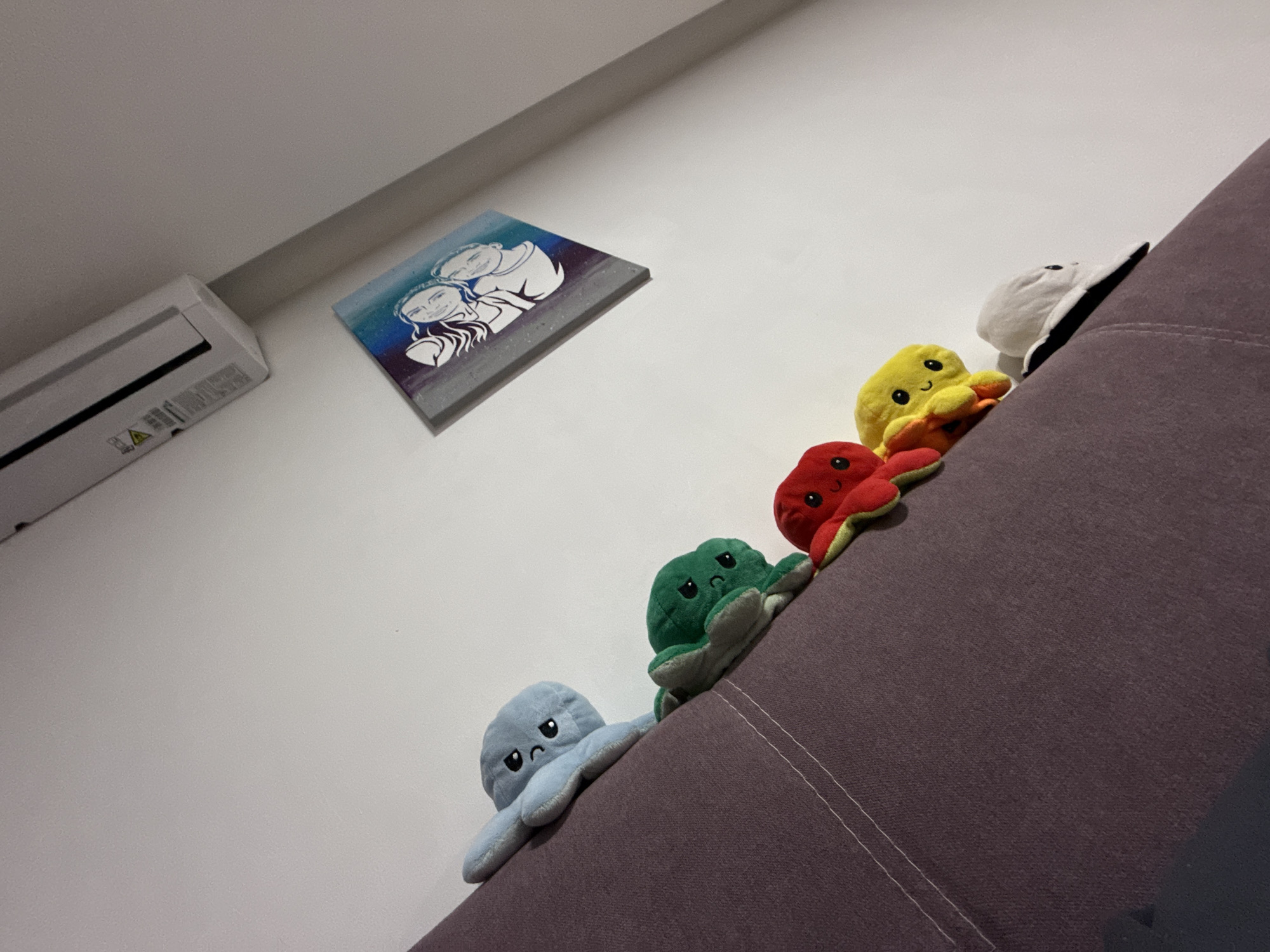 The image shows a low-angle view of a room interior. An air conditioning unit is mounted on a white wall, adjacent to a framed picture depicting two figures in a graphic style with blue accents. Arranged in a diagonal line on a purple upholstered surface, likely a sofa, are five plush octopus toys, each a different color (light blue, green, red, yellow, and white). The scene has a playful, domestic quality. The image provides insufficient information to determine a specific location in Plovdiv, Bulgaria.