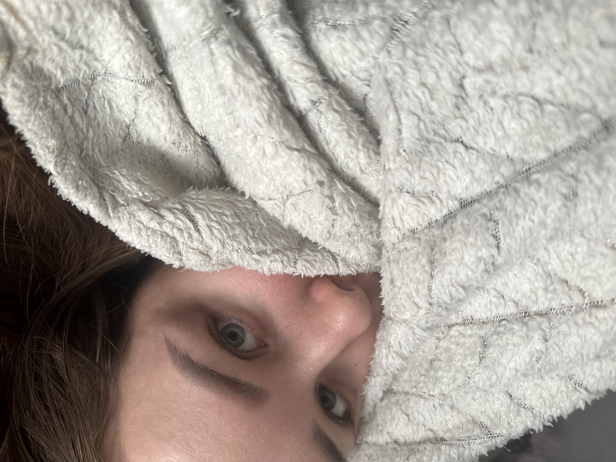 A close-up, off-center image shows a person's face mostly covered by a light gray, plush blanket with a quilted pattern. The person's eyes are visible, appearing light-colored. Brown hair is visible on the left side of the frame. The lower portion of the face, including the nose, is revealed just above the edge of the blanket. The location is not discernible from the image, so it is impossible to determine if it is in Chaikovsky, Russia.
