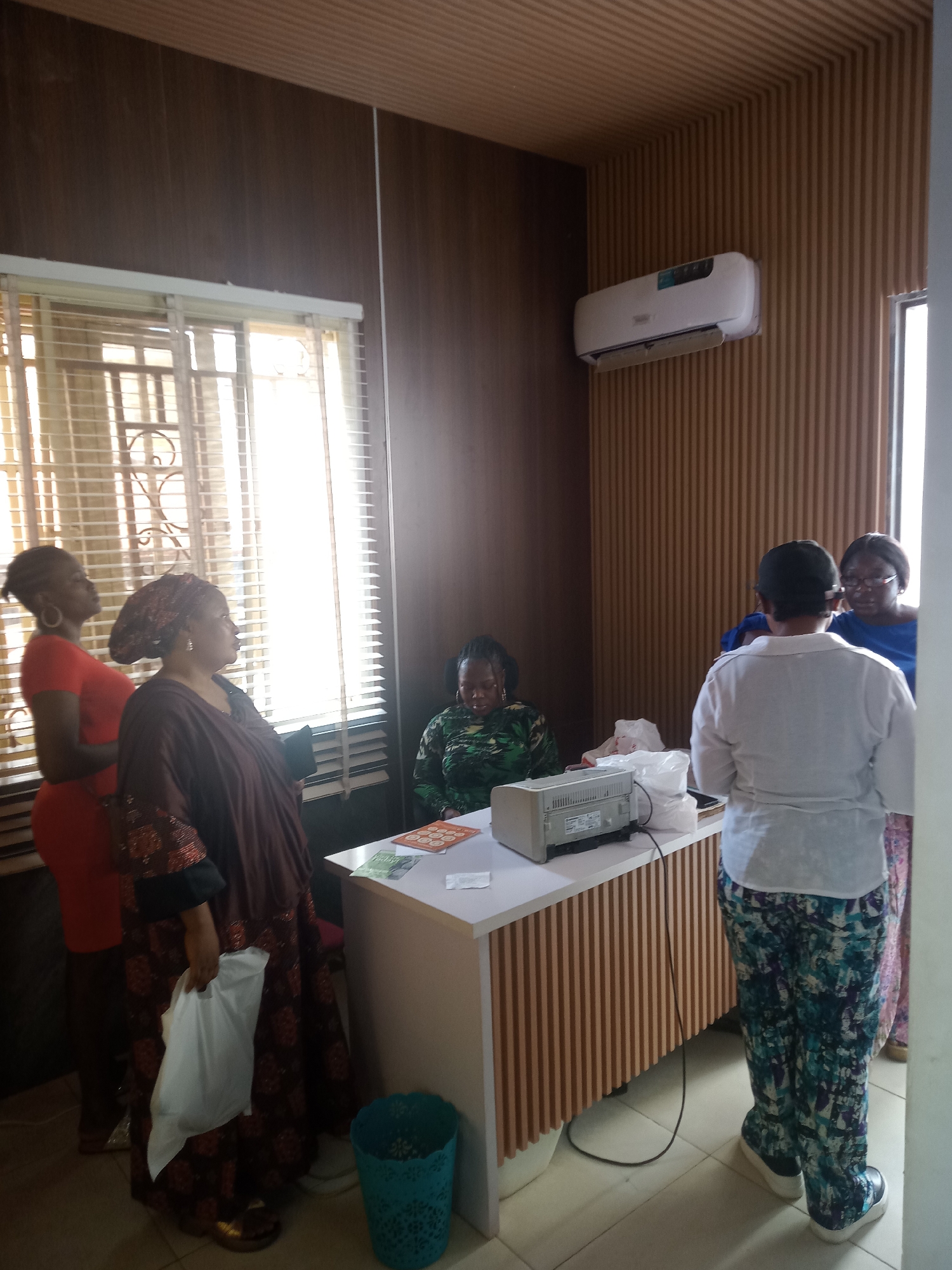 The image shows an indoor scene in what appears to be a small office setting, possibly located in Gwarinpa, Nigeria. There are five women present. Two women stand to the left, near a window with blinds, with one wearing a red dress and the other in a brown patterned dress and headwrap, holding a white plastic bag. A third woman sits behind a white desk, wearing a green patterned top. A fourth woman, wearing a white top and colorful pants, stands with her back to the camera. A fifth woman with glasses is seen in the background, wearing a blue top. On the desk, there is a gray printer, some white plastic bags or containers, and some pamphlets or brochures. A turquoise waste bin is located near the woman in the brown dress. The walls are decorated with horizontal wooden paneling, and an air conditioning unit is mounted high on the wall. The overall lighting is bright, indicating natural light entering through the window.