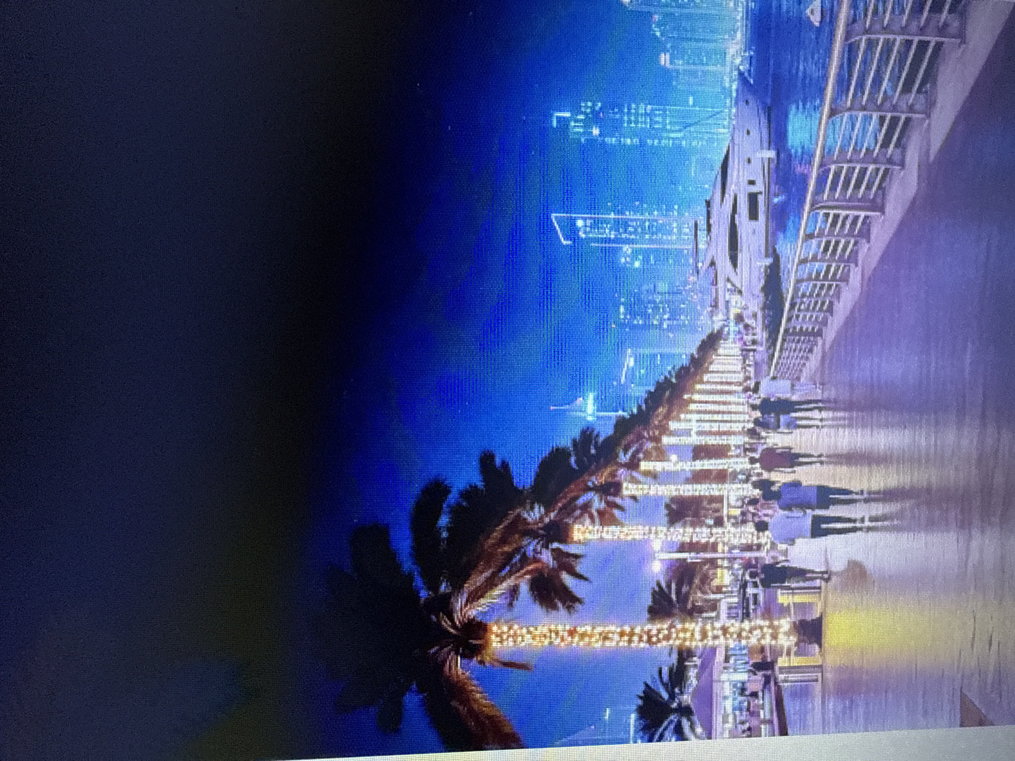 The image depicts a nighttime scene along a waterfront promenade. Several people are walking on a paved walkway lined with palm trees adorned with festive lights. The promenade overlooks a body of water where a large white yacht is docked. In the background, a city skyline with numerous illuminated skyscrapers creates a vibrant urban backdrop. The sky is dark blue, suggesting it is evening or night. There is no indication that this scene is located in Pushkino, Russia.