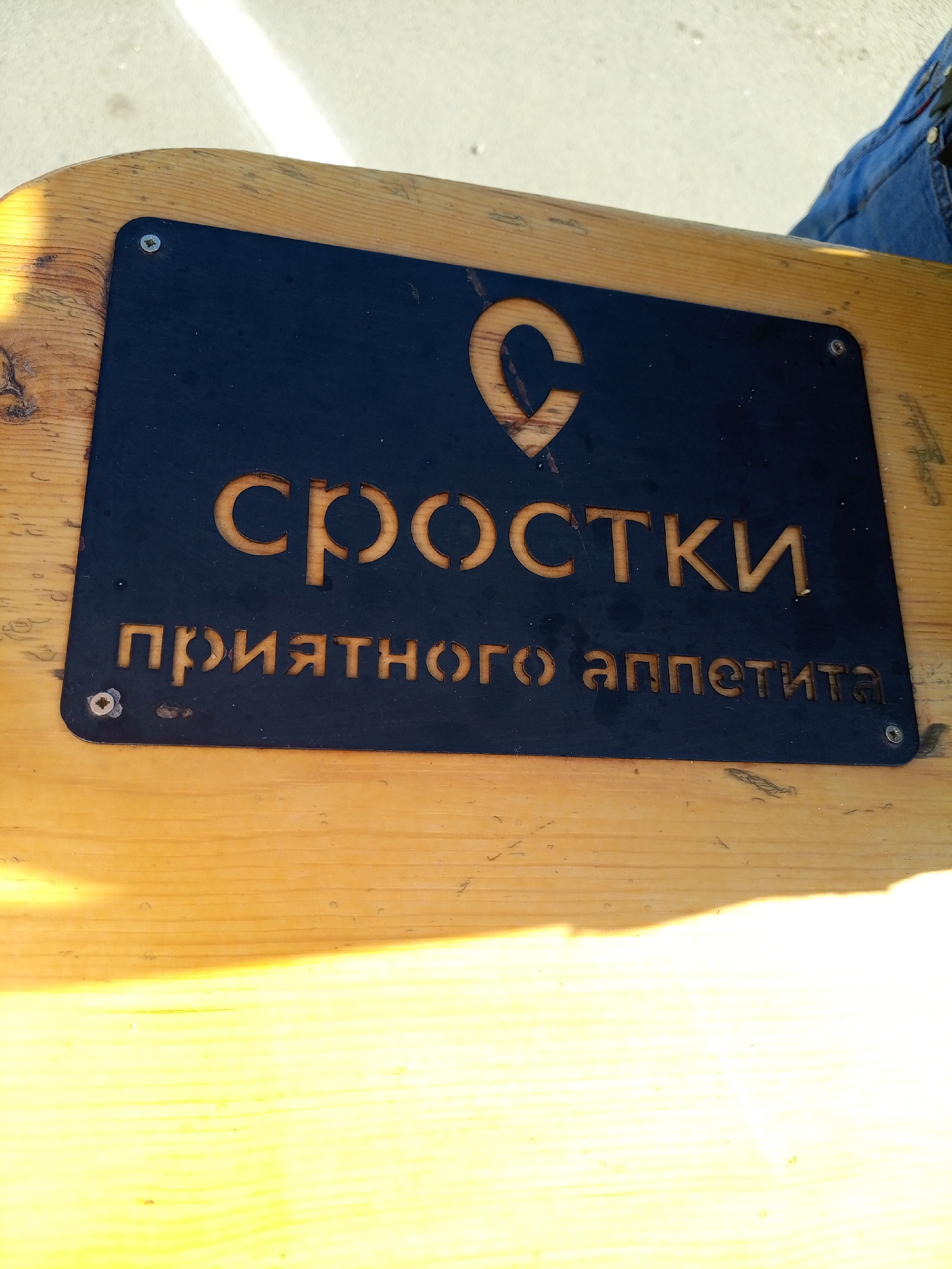 A close-up, eye-level shot shows a weathered wooden table with a black metal sign attached. The sign features a stylized orange letter "C" at the top, resembling a location pin. Below it, the word "СРОСТКИ" (Srostki) is carved in orange, followed by the phrase "ПРИЯТНОГО АППЕТИТА" (Bon Appétit). The metal is fastened to the wood with visible screws. The background is out of focus, showing a bright, sunlit area and a person's blue denim jeans. The scene appears to be outdoors, possibly in a public seating area.