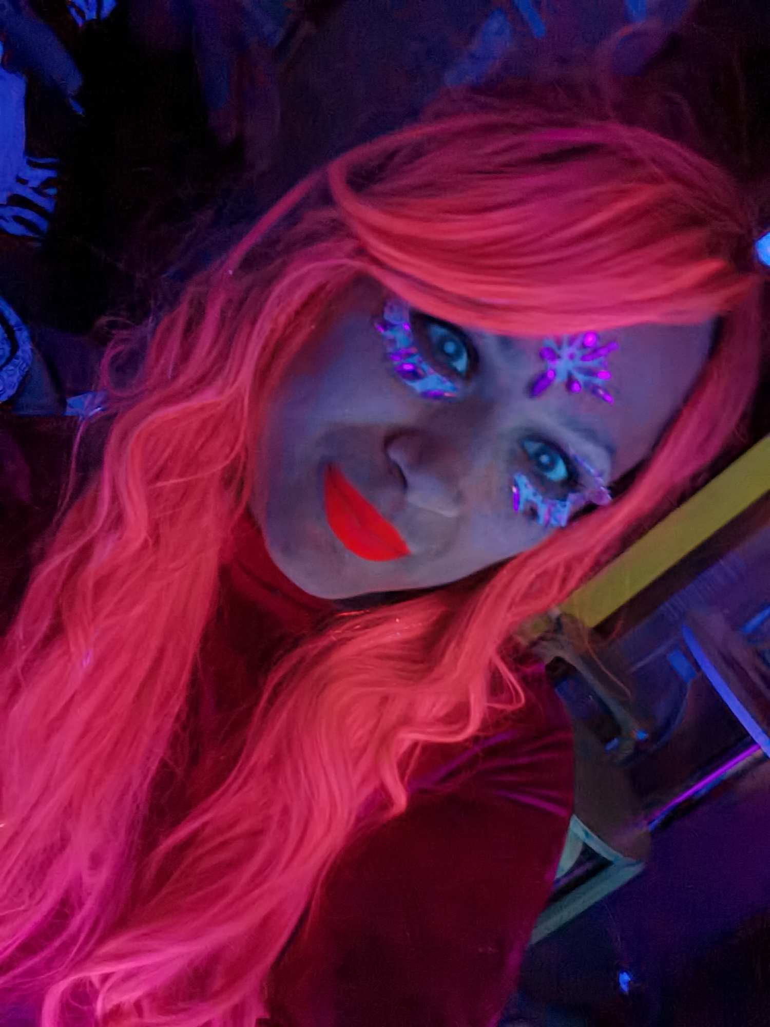 A close-up, tilted selfie shows a person with pink hair, lipstick, and gemstone stickers on their face. The person is wearing a red velvet turtleneck and is illuminated by ultraviolet (UV) light, causing their hair, makeup, and gemstones to fluoresce. The person's skin appears blue due to the UV lighting. The location appears to be a nightclub in the city of Chaikovsky, Russia. The background features dark and partially visible furniture.