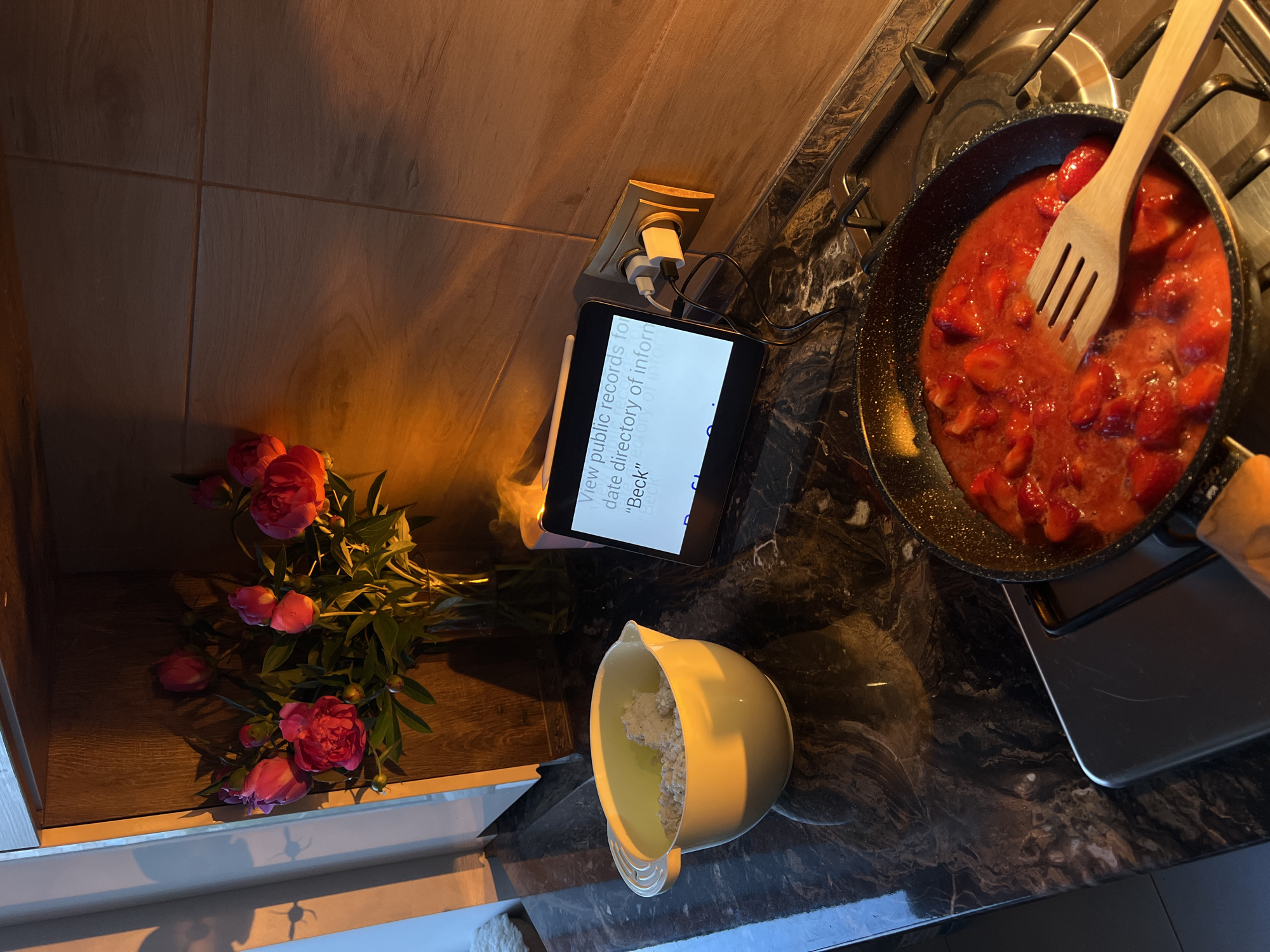The image displays a kitchen counter scene in Ternopil, Ukraine, illuminated by strong, warm, low-angle sunlight from the left, casting distinct shadows. No individuals are visible.
On the left, a light yellow mixing bowl containing a white, granular substance, consistent with cottage cheese, rests on a dark, glossy, mottled countertop. Directly behind the bowl, a clear glass vase holds a bouquet of vibrant pink and red peonies, positioned on a wooden shelf or cabinet surface. A window frame is partially visible to the far left, where the sunlight originates, casting a prominent spider-like shadow onto an adjacent white wall.
Towards the center-right, an electronic tablet, possibly an iPad, stands vertically, displaying partially legible text including "View public records to date directory of info 'Beck'". The tablet is connected by a white charging cable to a multi-port wall adapter plugged into a standard wall outlet. Beside the tablet, a small, cylindrical object, likely an essential oil diffuser or a candle, emits a faint wisp of vapor or smoke. The wall behind this area is tiled with light brown, wood-grain patterned tiles.
In the foreground, on a gas stovetop, a black frying pan contains a simmering preparation of sliced red strawberries in a sauce-like consistency. A wooden spatula with three tines rests within the cooking strawberries. The metal grates and burners of the stovetop are clearly visible beneath the pan.