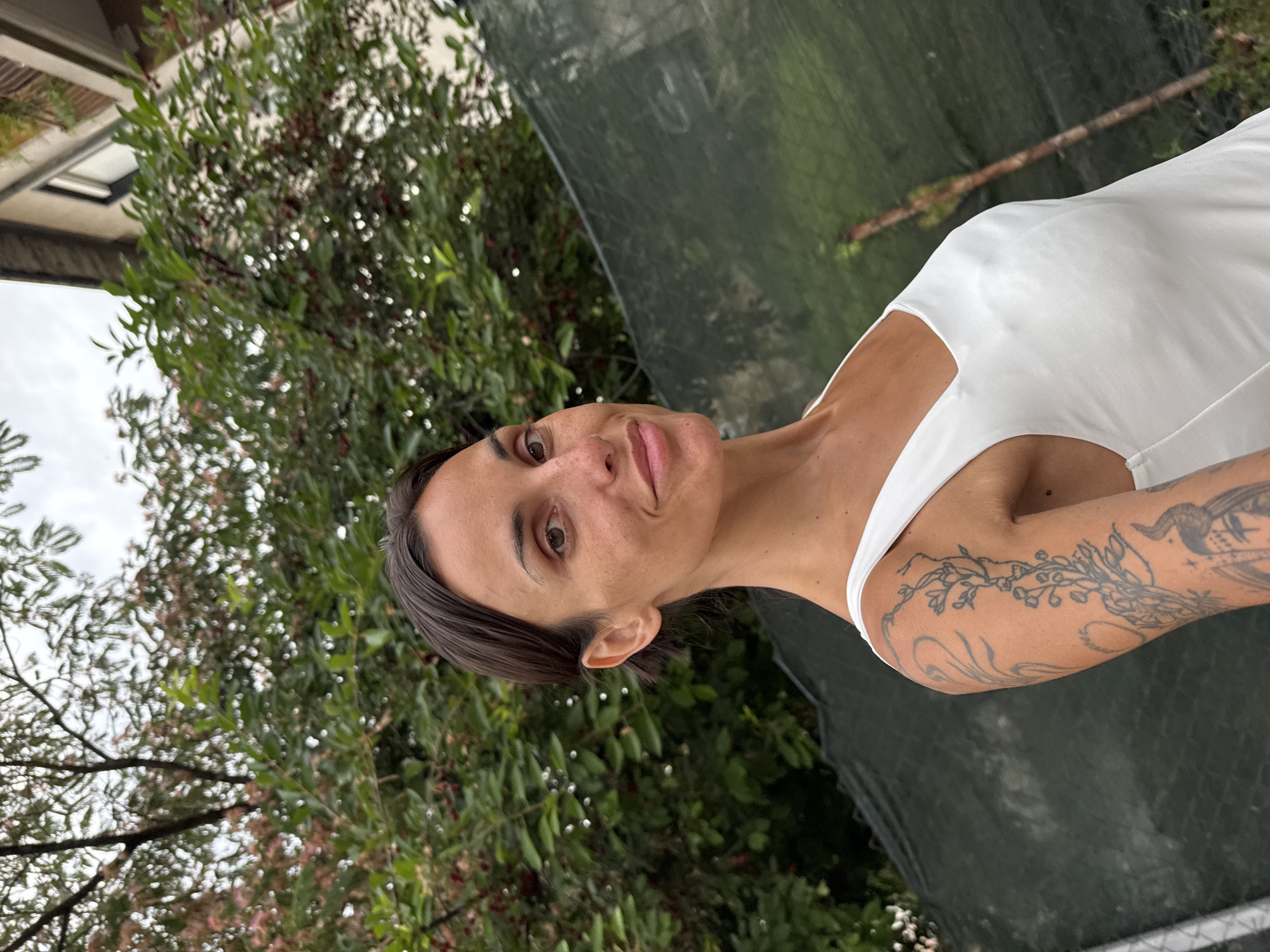 A woman with dark hair and fair skin, wearing a white tank top, poses for a selfie outdoors. She has freckles and visible tattoos on her left arm. The background features a dense green tree with some red berries, a dark green fence, and a building with a balcony. The scene appears to be a residential area. The image is taken in the city of Pleven, Bulgaria.