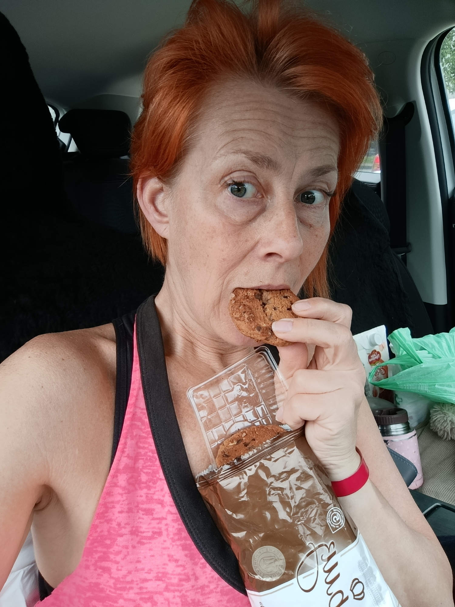 A woman with bright, coppery-red hair and fair skin is depicted from the chest up, seemingly inside a car. She is wearing a pink and black athletic tank top. Her gaze is directed towards the viewer with wide, expressive eyes, and her mouth is open as she takes a bite from a chocolate chip cookie held in her right hand. Her left hand holds a clear plastic container, which is part of a larger brown package of cookies, exposing more cookies inside.
The setting is the interior of a car, with a black car seat and headrest visible behind her to the left. On her right, through a car window, glimpses of green foliage and a brighter outdoor area are visible, suggesting it is daytime. Clutter on the passenger seat or a console next to her includes a crinkled green plastic bag, a small white carton with some Cyrillic text on it (partially obscured), and a pink-patterned thermos or bottle. A red wristband is visible on her left wrist.
The primary activity is the woman eating a cookie. Her expression suggests she might have been caught in the act or is playfully acknowledging the camera. On the brown cookie packaging, the word "Pure" (or a similar word with a script font) is visible, along with some circular logos and other small text. The overall scene conveys a casual, on-the-go moment, possibly in Moscow, Russia.