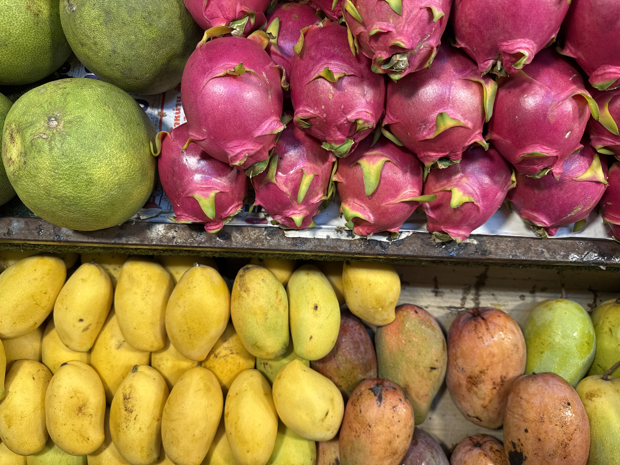 A close-up, overhead, vertical shot shows a selection of fresh fruit displayed in what appears to be a market stall. On the left side of the frame, a large quantity of yellow and some reddish-orange mangoes are piled high. The mangoes vary in ripeness and size. To the right of the mangoes, separated by a metal grate, are bright pink dragon fruits with vibrant green leafy scales. Above the dragon fruits, and partially visible behind them, are several large, pale green pomelos. The background is indistinct, but appears to be composed of newspaper or paper liners within the stalls. The image captures the colors and textures of the fruit, suggesting a vibrant, fresh produce offering in Rawai, Thailand. There are no people or actions depicted in the image.