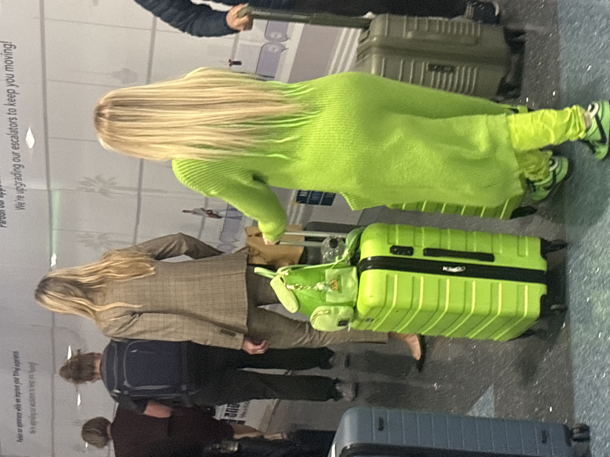 At an airport, two women with blonde hair stand among other travelers with luggage. One woman is wearing a lime green sweater and matching pants and shoes. The other is wearing a tan blazer and pants. The airport is brightly lit and there is a sign in the background that reads, "Pardon our appearance while we upgrade your travel experience."