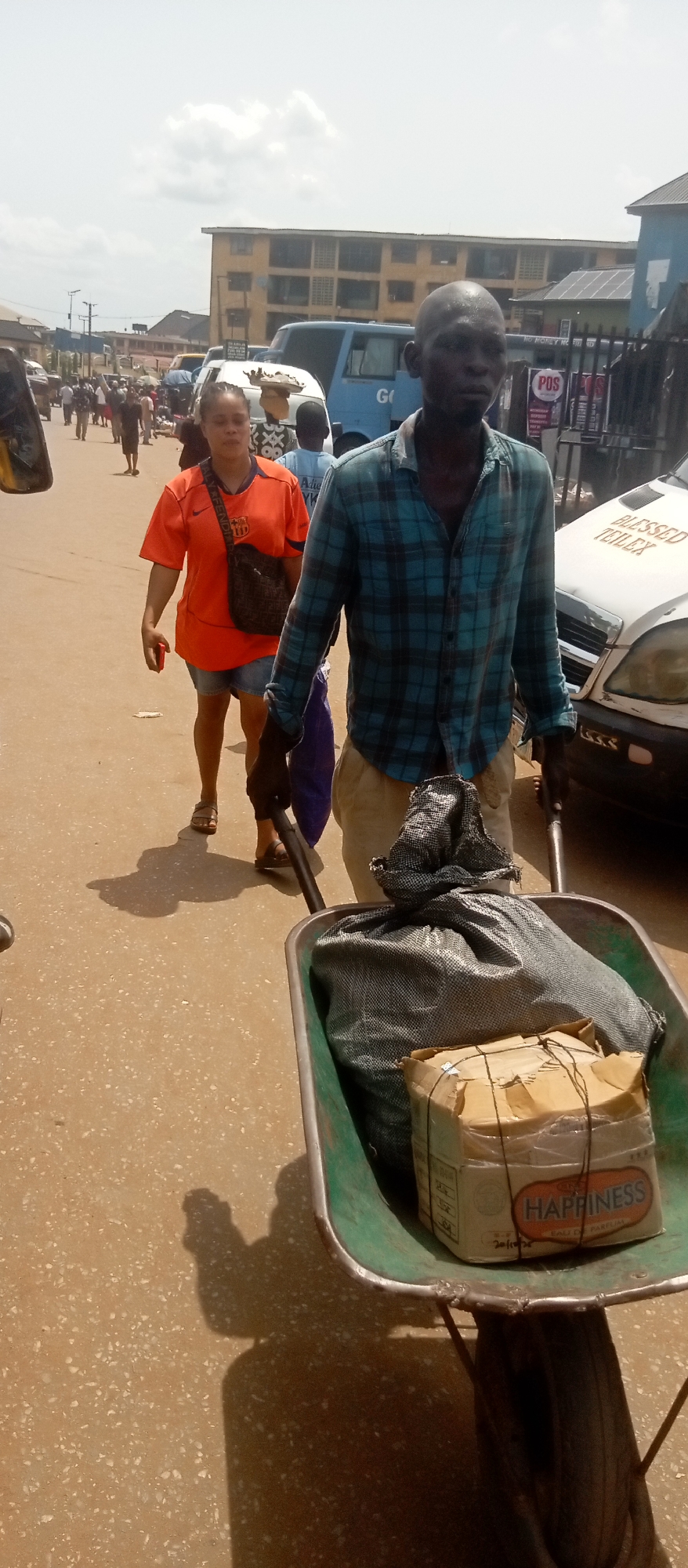 In Onitsha, Nigeria, a man pulls a green wheelbarrow down a sunny street. The wheelbarrow contains a black woven sack and a taped cardboard box labeled "HAPPINESS". The man wears an unbuttoned plaid shirt, khaki pants, and gloves. A woman in an orange Barcelona shirt, denim shorts, and sandals walks beside him, carrying a purse. Other pedestrians and vehicles fill the street. A white car with the text "BLESSED TEILEX" is parked nearby. POS signage is visible on a building in the background.