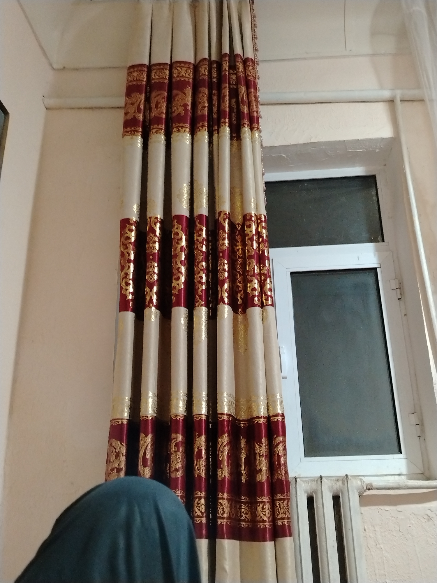 In Nukus, Uzbekistan, the image captures an interior scene with a focus on a window and curtains. A section of a white radiator is visible beneath the window, suggesting it is likely a room in a residential building. The window itself is framed in white and has a dark, possibly tinted or frosted, glass pane. To the left of the window, a set of heavy curtains with a cream base and a repeating pattern of dark red and gold ornamentation are drawn. The pattern appears to be floral or a decorative motif. In the foreground, the top of a person's head is visible, covered with a dark teal fabric, obscuring their face and most of their body. The person is positioned to the left of the frame, partially behind the curtains. The wall behind the curtains and the window appears to be a light, neutral color. The overall lighting suggests an indoor setting, possibly during daytime or when artificial lights are on.