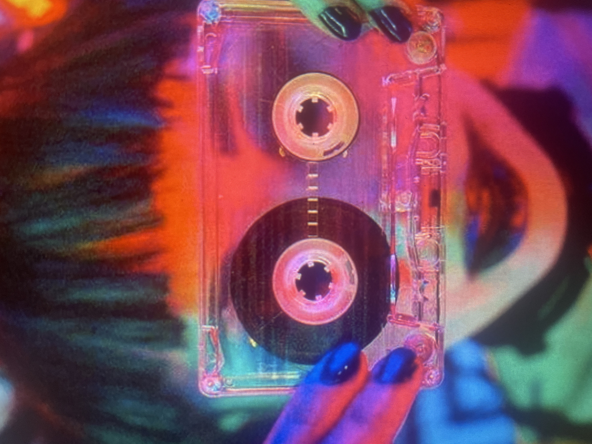 A close-up, head-and-shoulders shot shows a person holding a clear cassette tape in the foreground. The person's hands, with dark nail polish, are visible on either side of the tape. The tape's spools are visible, and the tape itself is transparent. The person's face is partially obscured by the tape and out of focus, but their lips are visible, painted a dark color. The background is a blur of vibrant, neon colors, predominantly reds, blues, and greens, suggesting a dimly lit, possibly urban environment. The image has a grainy, lo-fi aesthetic, possibly due to low light conditions or intentional artistic choice. No specific location in the city of Unknown, Unknown is identifiable.