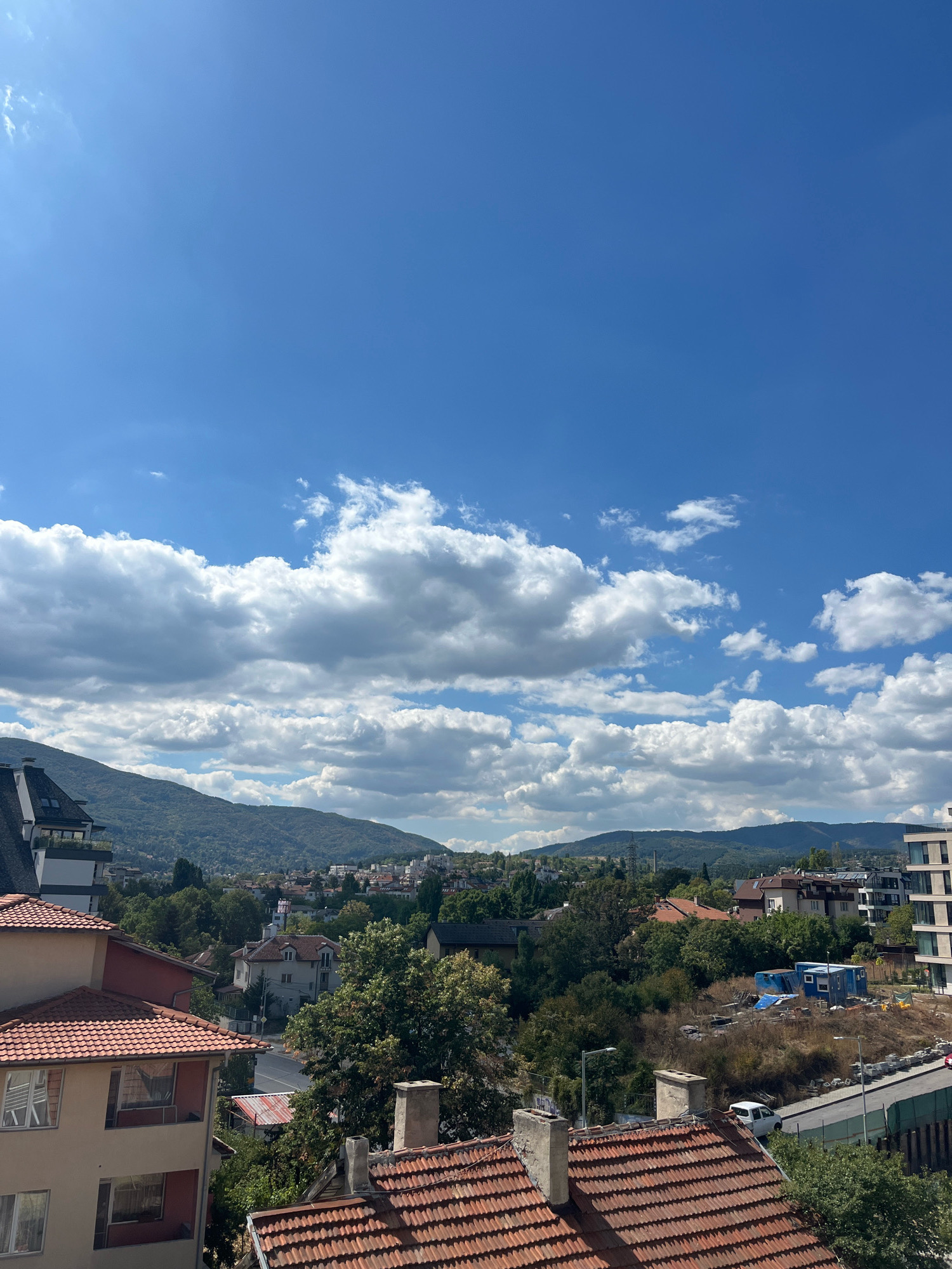 The image captures an elevated view of a cityscape under a bright, partly cloudy sky. The scene depicts a dense urban environment with a mix of architectural styles, from older residential buildings with red tile roofs to modern multi-story structures. Lush green trees are interspersed throughout the city, and a backdrop of rolling, tree-covered hills suggests the city is situated near a mountainous region. In the middle ground, there appears to be a construction site with temporary blue structures and materials. The sky is predominantly a vibrant blue, with scattered cumulus clouds casting soft shadows. The overall impression is of a sunny day in a European city, likely Sofia, Bulgaria, given the architectural and landscape cues often associated with the city. No people are visible in the image.