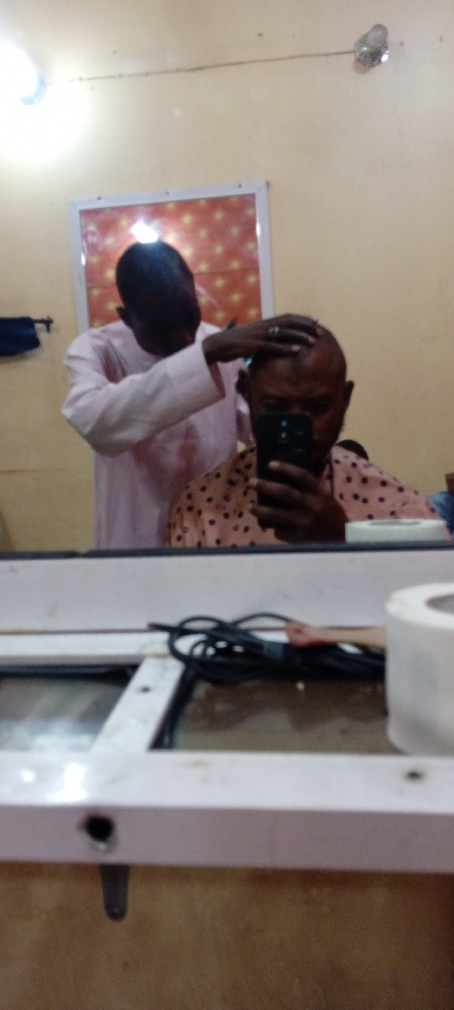 The image captures a scene inside what appears to be a small barbershop. A dark-skinned man in a light pink robe is standing behind another dark-skinned man, who is seated and holding a smartphone, presumably taking a selfie in the mirror. The standing man is touching the seated man's head, indicating he might be cutting or styling the hair. The seated man is wearing a salmon-colored shirt with dark polka dots. The mirror reflects the image. A white frame surrounds the mirror, and an electrical cord is visible in the foreground on what looks like a shelf or table. A roll of white material, possibly paper or cloth, is also present on the shelf. On the wall, there's a framed picture. The lighting in the room is soft, with a light fixture visible in the upper left corner of the frame. The precise location and city are not discernible from the image.