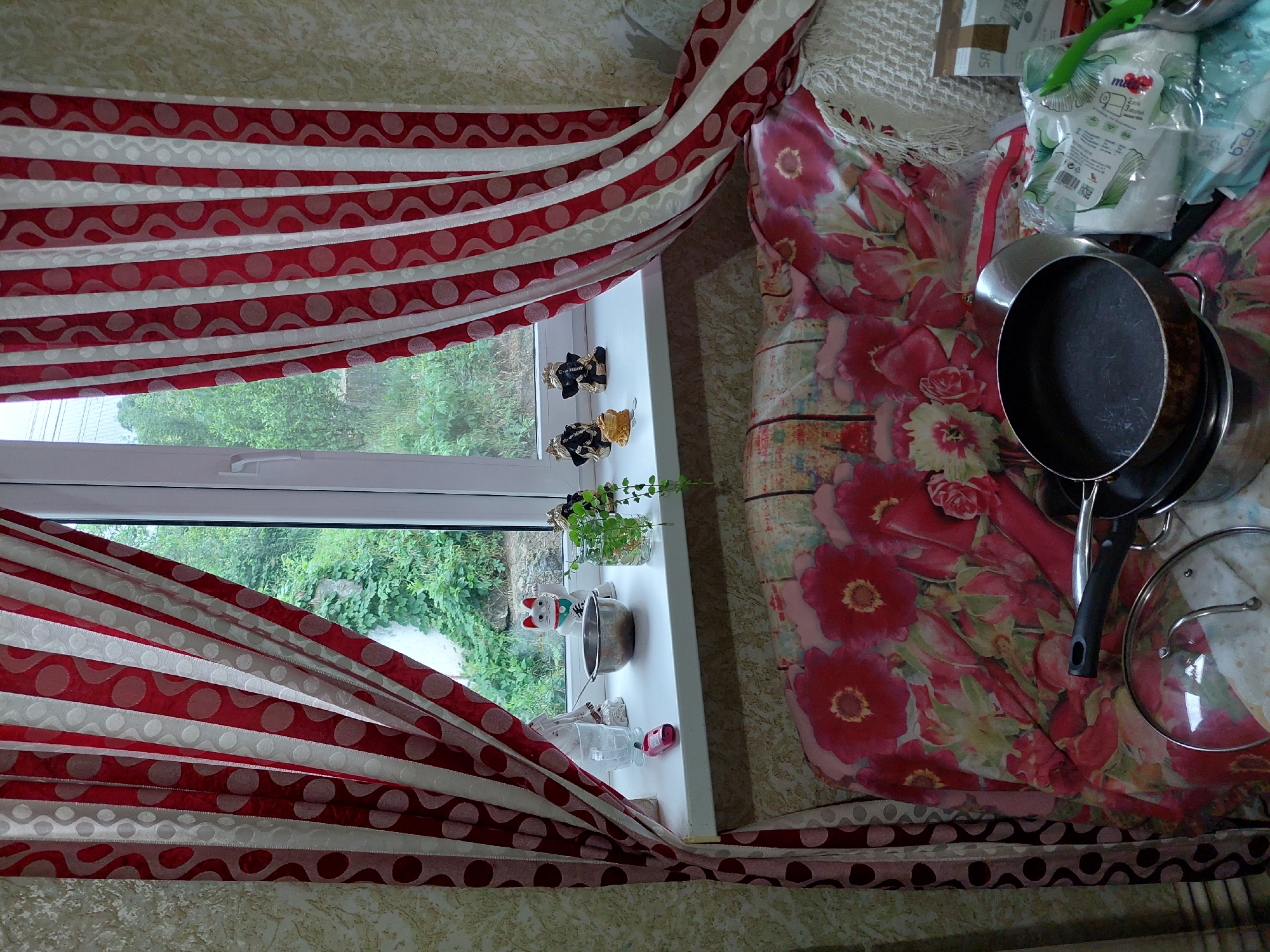 This scene is situated in a domestic interior in the city of Orhei, Moldova. The image captures a cluttered window area and the space directly beneath it, indicating a lived-in environment.
The central feature is a white-framed window, providing a view of dense green foliage, including trees, bushes, and vines, with a light-colored wall partially visible behind the greenery. Two curtain panels flank the window, pulled back towards the walls. These curtains are red and white, featuring vertical stripes. The red stripes are adorned with a pattern of white circles, while the white stripes exhibit a subtle wavy or leafy pattern.
On the white windowsill, arranged from left to right, are: a partially obscured white figurine, an empty clear drinking glass, a small pink electronic device, a small metallic bowl, a white ceramic Maneki-neko (waving cat) figurine with a red collar and green bell/pendant, a clear glass jar containing a small green plant (possibly a vine) rooted in water, two dark figurines with gold crowns (resembling Ganesha), and a small golden animal figurine (possibly a frog or toad) positioned in front of two small coins.
Below the windowsill, a piece of furniture, likely a sofa or bed, is covered with a vibrant textile featuring a large floral pattern of red and pink flowers and green leaves. On the right side of this covered furniture, a white crocheted or knitted blanket is visible. Various household objects are arranged on this surface: a clear glass lid with a metal handle, a dark-colored frying pan with a black handle resting on another metallic pot, additional metal cookware beneath the frying pan, a stack of plastic-packaged hygiene products (including packages labeled "baby" and containing wet wipes), a green plastic utensil (possibly a spatula or spoon), and a partially visible cardboard box on the far right, marked "S A R S". No people are present or engaged in direct actions within the frame.