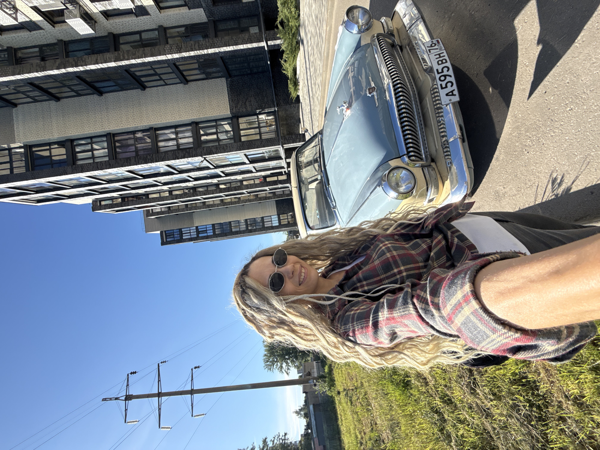 The image, taken in Rusanovo, Russia, presents a vibrant outdoor scene on a clear, sunny day. The focal point is a young woman taking a selfie, positioned to the right of a vintage car.
The woman has long, wavy blonde hair, possibly styled with dreadlocks or braids, and wears round sunglasses. She is dressed in a red, black, and white plaid flannel shirt over a white undershirt, and displays a cheerful, open-mouthed smile directed at the camera. Her right arm is extended, indicating she is holding the camera for the photograph.
To the woman's left, a classic light blue and beige car is parked on asphalt pavement. The vehicle is notable for its prominent chrome grille, shiny chrome bumper, and round headlights. A Russian license plate, "A 595 BH 76 RUS," is affixed to its front.
The background features a blend of modern architecture and natural elements. To the left of the car, tall, contemporary buildings with dark windows and brick facades rise. Further in the distance, a large utility pole with power lines stands against a bright, cloudless blue sky. The foreground to the right consists of a sunlit grassy area, while asphalt covers the ground where the car is parked. Strong shadows from the car, person, and buildings are cast onto the pavement, reinforcing the impression of a bright, high-sun setting. The overall atmosphere of the image is pleasant and relaxed.