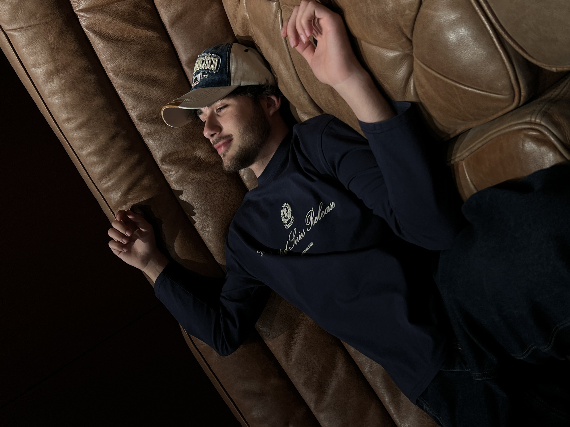 A young man is lying on a brown leather couch. He is wearing a two-tone baseball cap, a long-sleeved navy blue shirt with an emblem on the chest, and dark jeans. The location is not discernible from the image.