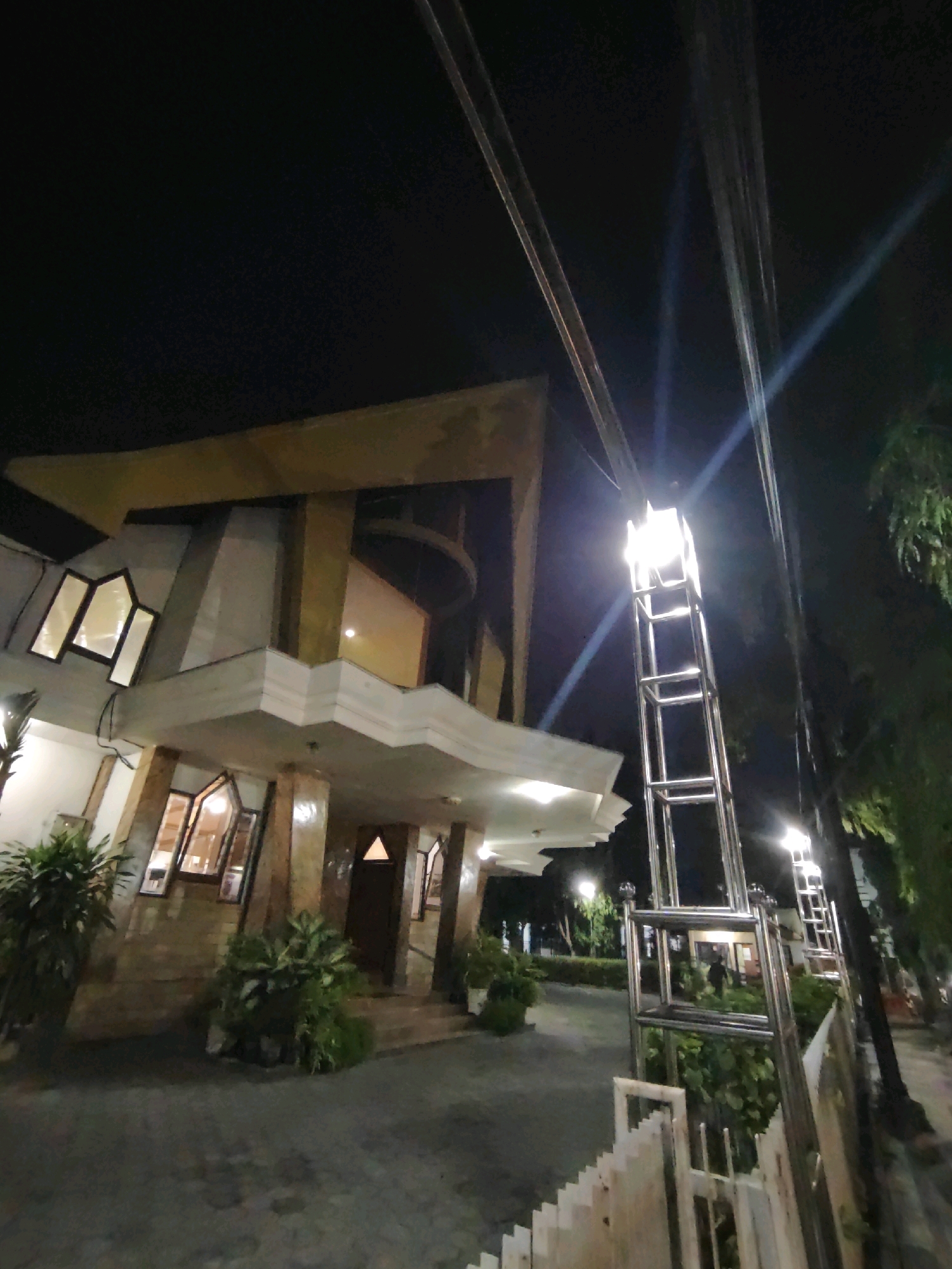 The image depicts a nighttime scene of a building, likely a church or a similar institution, in Kediri City, Indonesia. The building features modern architecture with geometric shapes and prominent windows that are illuminated from within. A tall, metallic lamp post stands prominently in the foreground, casting bright light and creating lens flare effects. Several utility poles and wires are visible against the dark sky. Lush green plants are strategically placed around the building and in planters, adding a touch of nature to the urban environment. The ground appears to be paved, possibly with cobblestones or concrete. The overall impression is one of a well-lit, contemporary structure set against the backdrop of a quiet, nocturnal urban landscape.