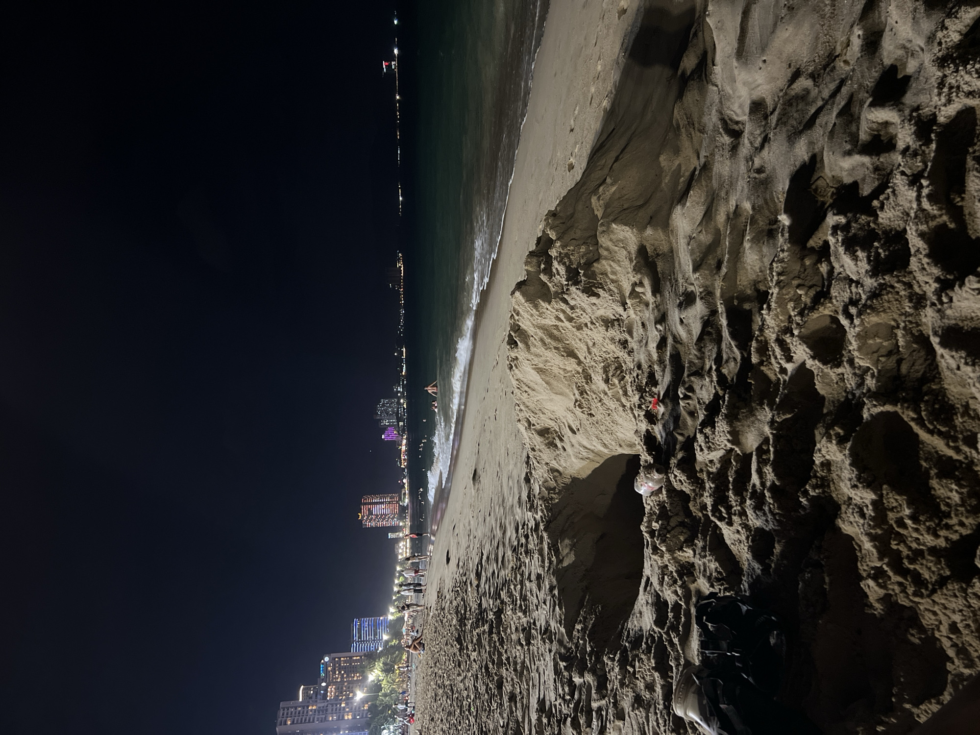 The image captures a night scene on a beach in Nha Trang, Vietnam, with the city skyline illuminated in the background. In the foreground, a large, deep hole has been dug into the sandy beach, with some discarded plastic bottles visible inside and near the hole. To the left of the hole, a black bag is placed on the sand. Further down the beach, a few people are seen walking and swimming in the ocean. The water is dark, reflecting the night sky, and waves are gently lapping at the shore. The city lights create a vibrant contrast against the dark night, highlighting the urban development along the coastline.