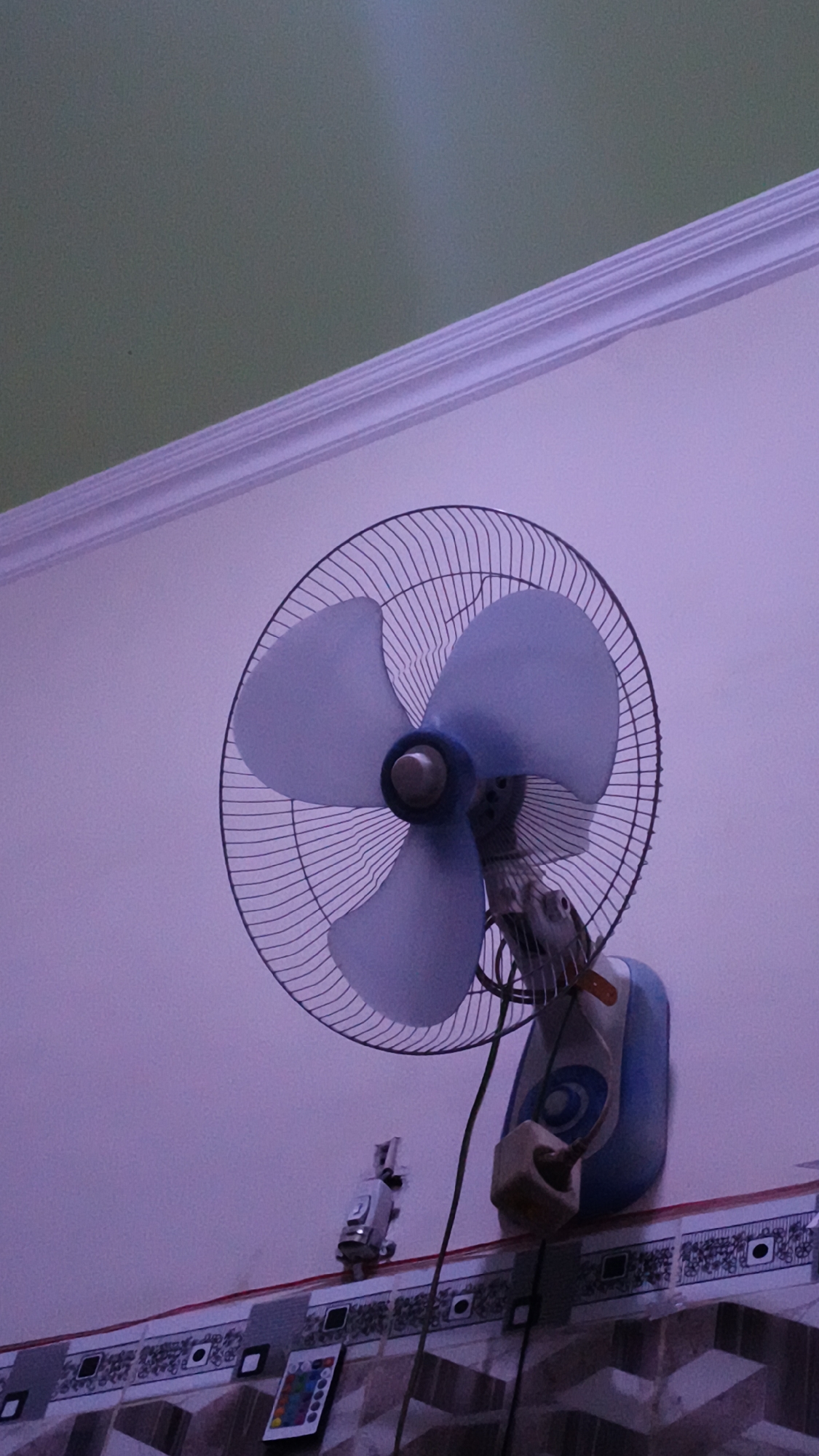 The image shows a wall-mounted electric fan with blue blades and a black wire grille. The fan is attached to a blue and white casing, which is mounted on a light purple wall. Above the wall is a cream-colored cornice and a greenish ceiling. Below the fan, on the wall, is a decorative border with a repeating pattern. To the left of the fan's casing is a small, silver-colored device with a clock face. Further down on the wall, near the decorative border, is a small, rectangular object with colorful buttons, possibly a remote control. The image appears to be taken indoors.