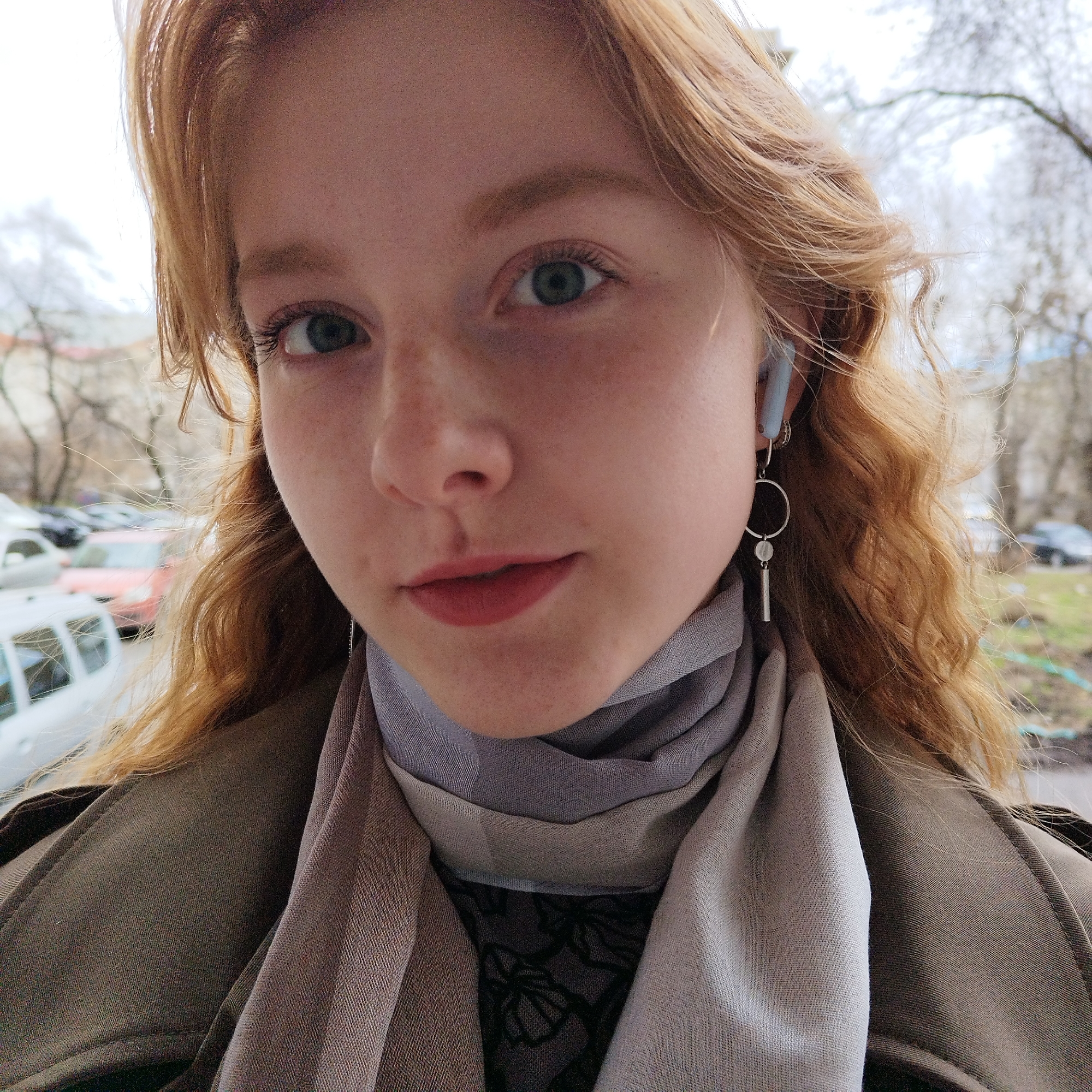 A young woman with long, reddish-blonde wavy hair is centered in the frame in an outdoor setting. She has fair skin with freckles, blue-green eyes, and is wearing light-colored lipstick. She is wearing a light gray and tan scarf wrapped around her neck, and a dark gray or olive green coat or jacket. Dangling silver earrings are visible on her left ear, and a small blue earbud is visible in her right ear. In the background are bare trees, a building, and several parked cars, indicating an urban environment, specifically Yekaterinburg, Russia.