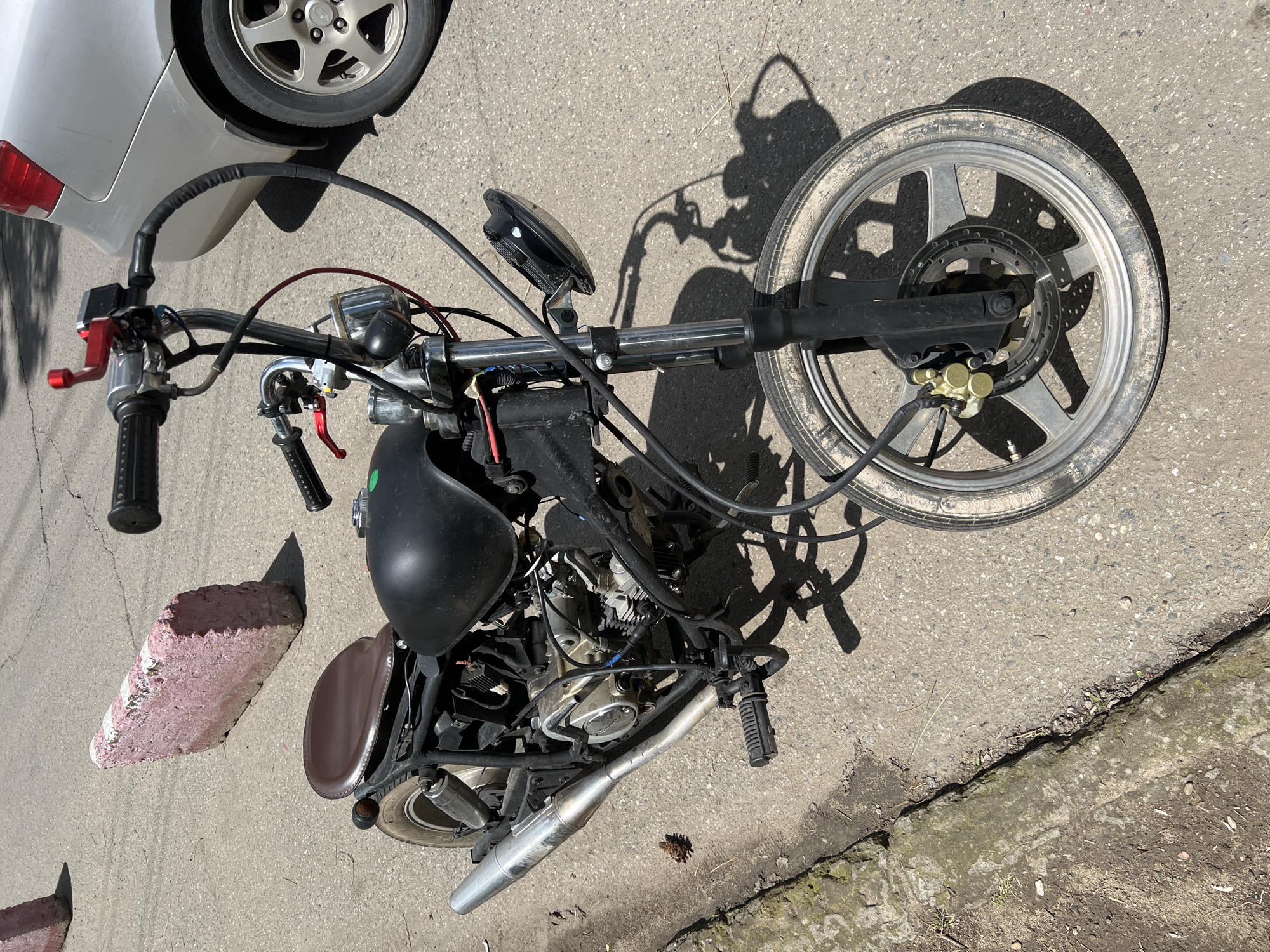 A custom motorcycle lies on its side on asphalt pavement. The motorcycle appears incomplete, with exposed wiring and a missing front fender. The location is suggested to be Krasnodar, Russia. A section of a silver car is visible at the top left. A pink brick or block lies on the left. The scene suggests a possible repair or modification project.