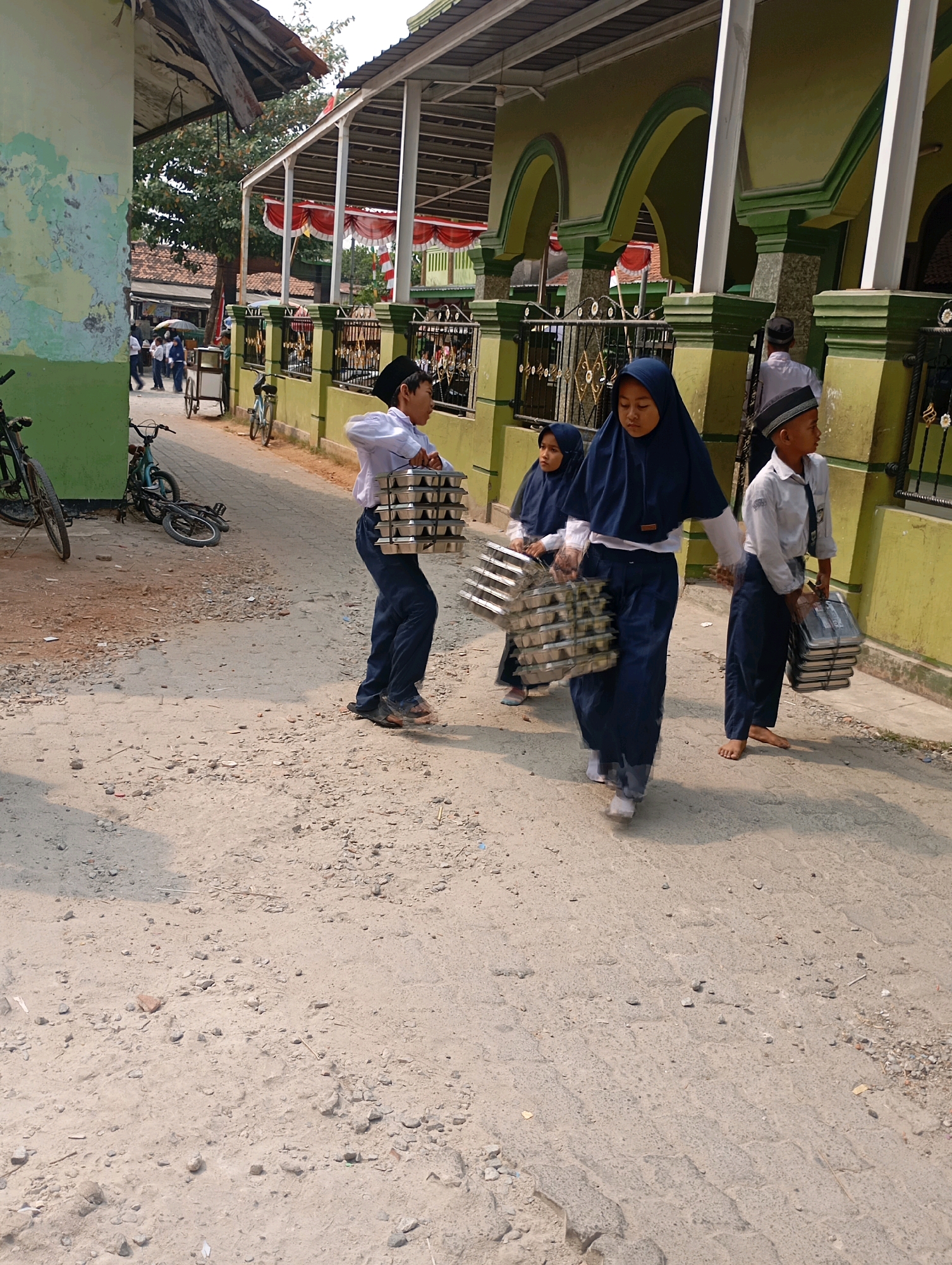 In Babelan, Indonesia, four young students, three boys and one girl, dressed in school uniforms, are walking on a dusty road beside a building with green and yellow pillars. The two boys in front are carrying stacks of metal trays. One boy, wearing a black cap and white shirt, walks barefoot. The girl, wearing a blue hijab and uniform, is also carrying trays. The third boy, in a white shirt and black cap, is walking behind them, carrying a stack of black trays. In the background, there are bicycles, a cart, and more students walking. The scene suggests a school or religious gathering, with decorations and banners visible on the building.