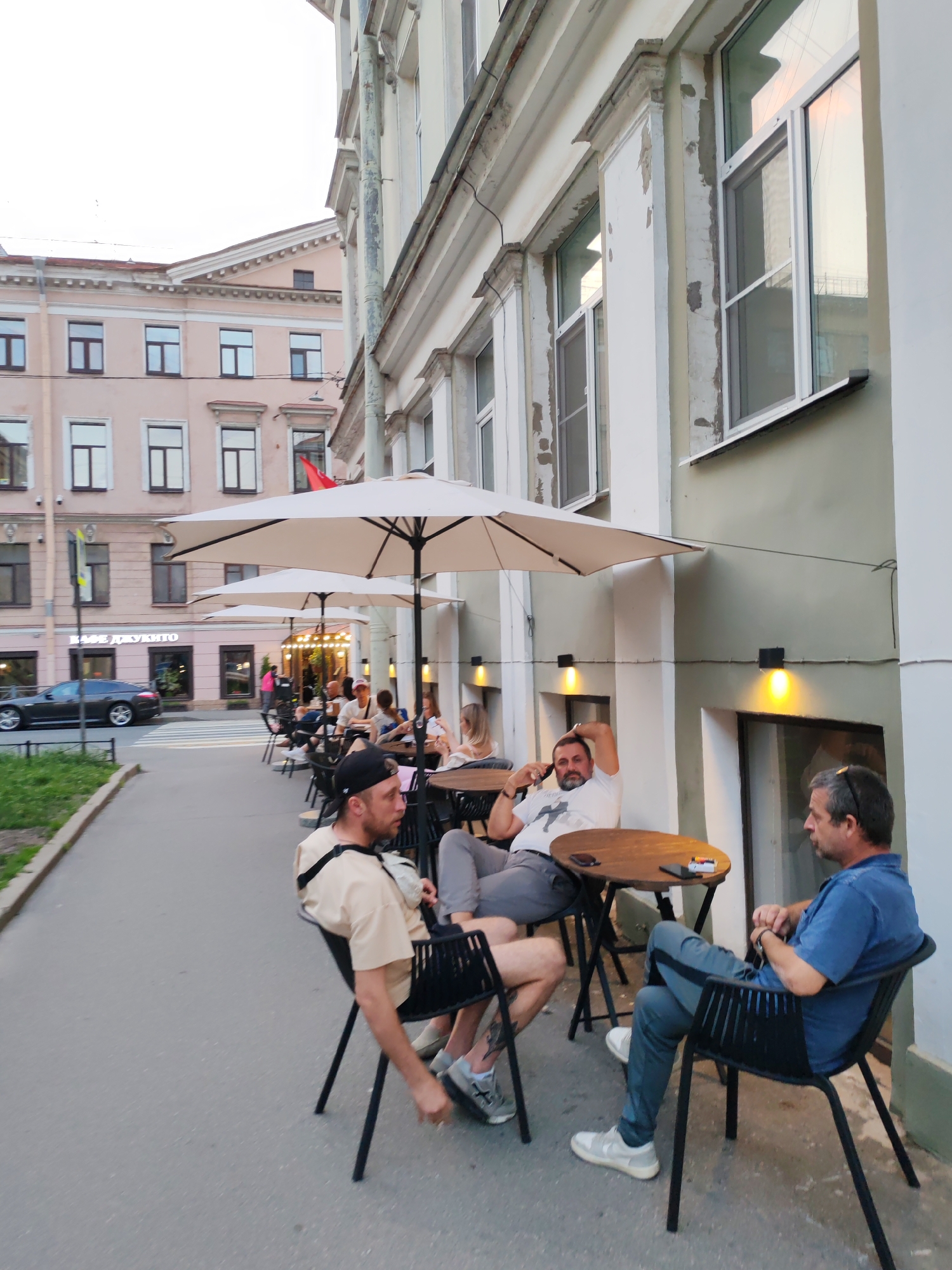 The image depicts an outdoor seating area of a cafe on a street in Saint Petersburg, Russia. Several people are seated at tables under large umbrellas. In the foreground, a man wearing a beige t-shirt and a black baseball cap is seated in a black chair, facing away from the camera. To his right, another man in a white t-shirt is leaning back in his chair with his arm behind his head. Further to the right, a third man in a blue t-shirt is seated and looking to his left. Several other individuals are visible at tables in the background, some interacting with each other. The cafe appears to be situated in front of an old building with a textured facade and multiple windows. A dark-colored car is parked on the street to the left, and a pedestrian crossing is visible. The lighting suggests it is either early morning or late evening.