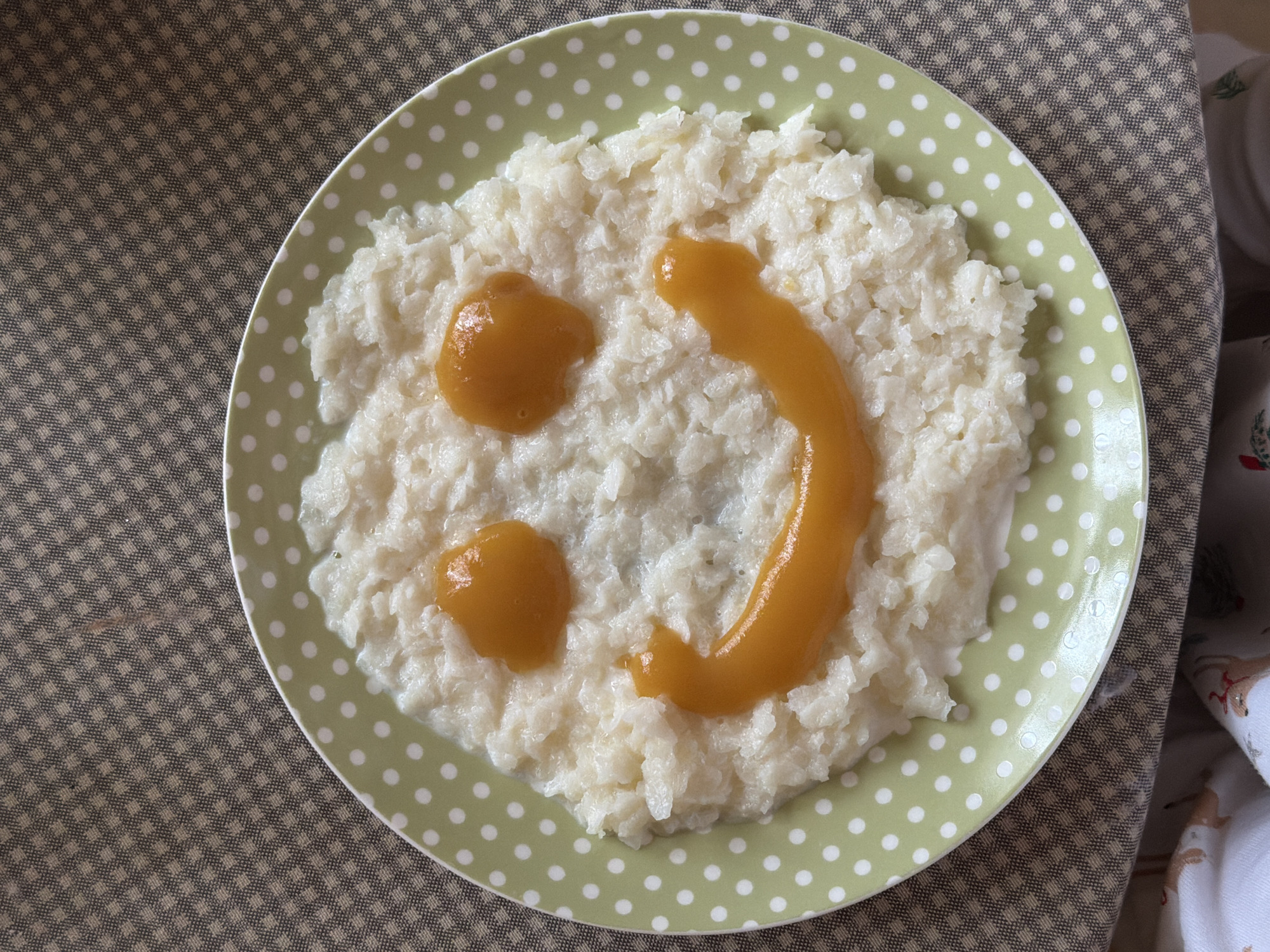 The image shows a plate of rice with what appears to be mango puree on top, arranged to resemble a smiley face. The plate is light green with white polka dots. The rice is white and slightly clumpy. The mango puree is a golden-orange color. The plate is placed on a brown and beige checkered surface. Part of a patterned white fabric is visible at the top-right corner of the image. The location (Khimki, Russia) is not identifiable from the content of the image.