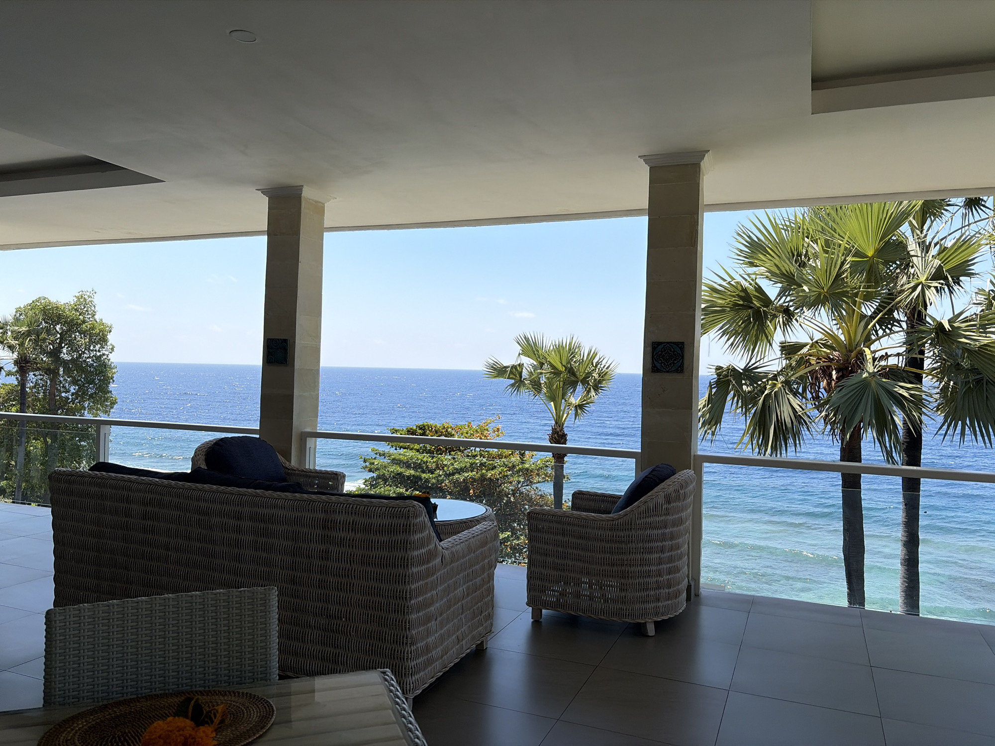 This is a wide-angle, eye-level shot of an outdoor seating area on a covered patio overlooking the ocean in Abang, Indonesia. The patio floor is tiled in a dark, glossy brown. Two large wicker sofas with dark blue cushions are positioned facing the ocean. To the right, a single wicker armchair with a dark blue cushion is also facing the ocean. In the foreground, part of a wicker chair and a small round table with a woven placemat and some orange and black decorative elements are visible. A glass railing separates the patio from the view of the ocean and a lush green landscape. Two palm trees are prominently featured in the mid-ground on the right, with their fronds extending over the railing. The ocean is a vibrant blue with gentle waves, and the sky is clear and blue. The scene conveys a sense of relaxation and tropical ambiance.