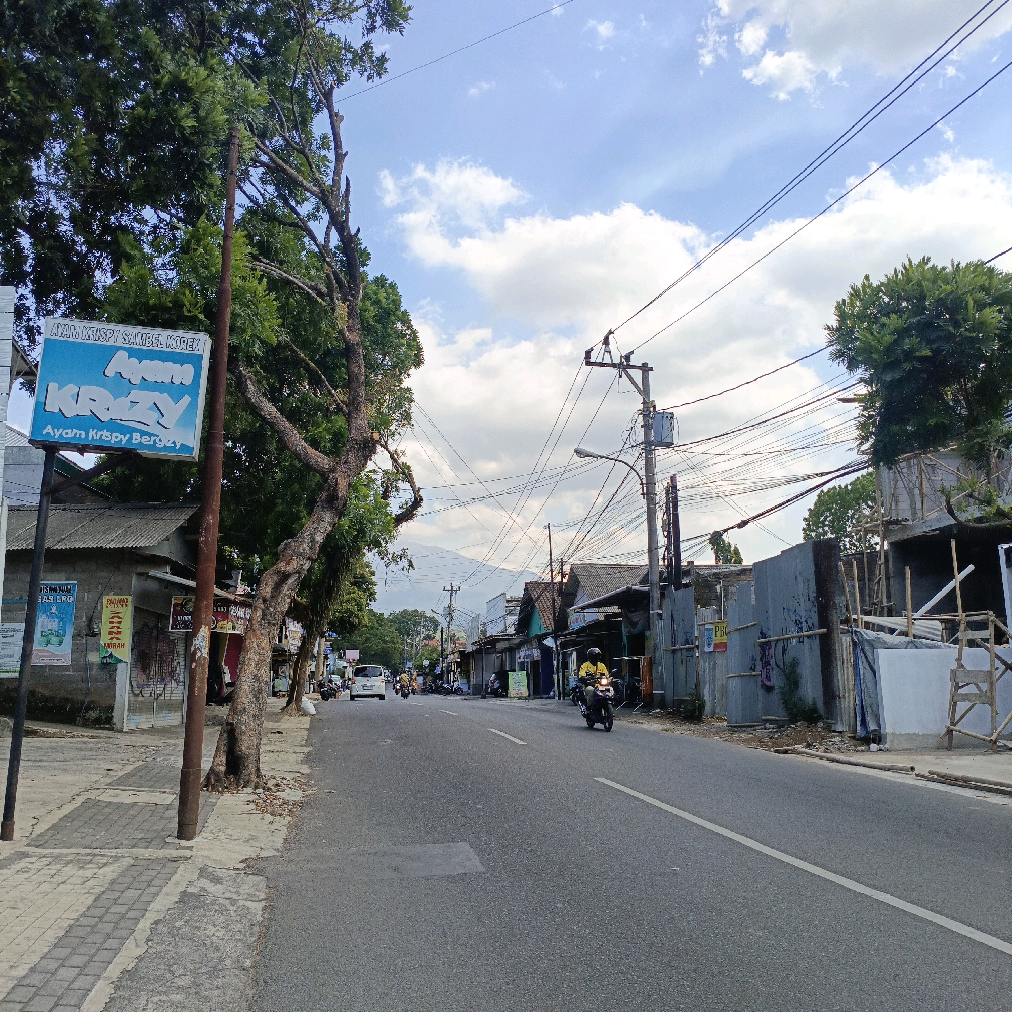 This is a street scene in Salatiga, Indonesia. The image is taken from the perspective of someone on the sidewalk looking down a road. On the left side of the road, there is a blue sign advertising "AYAM KRISPY SAMBEL KOREK" and "AYAM KRAZY". Next to the sign are trees and a building with a corrugated metal roof. Further down the street, there are more buildings and shops on both sides of the road. Several motorcycles and a white car are visible on the road. The sky is partly cloudy with a few white clouds against a light blue background. In the distance, a mountain range is faintly visible through the haze. The overall scene depicts a typical commercial street in an Indonesian city.