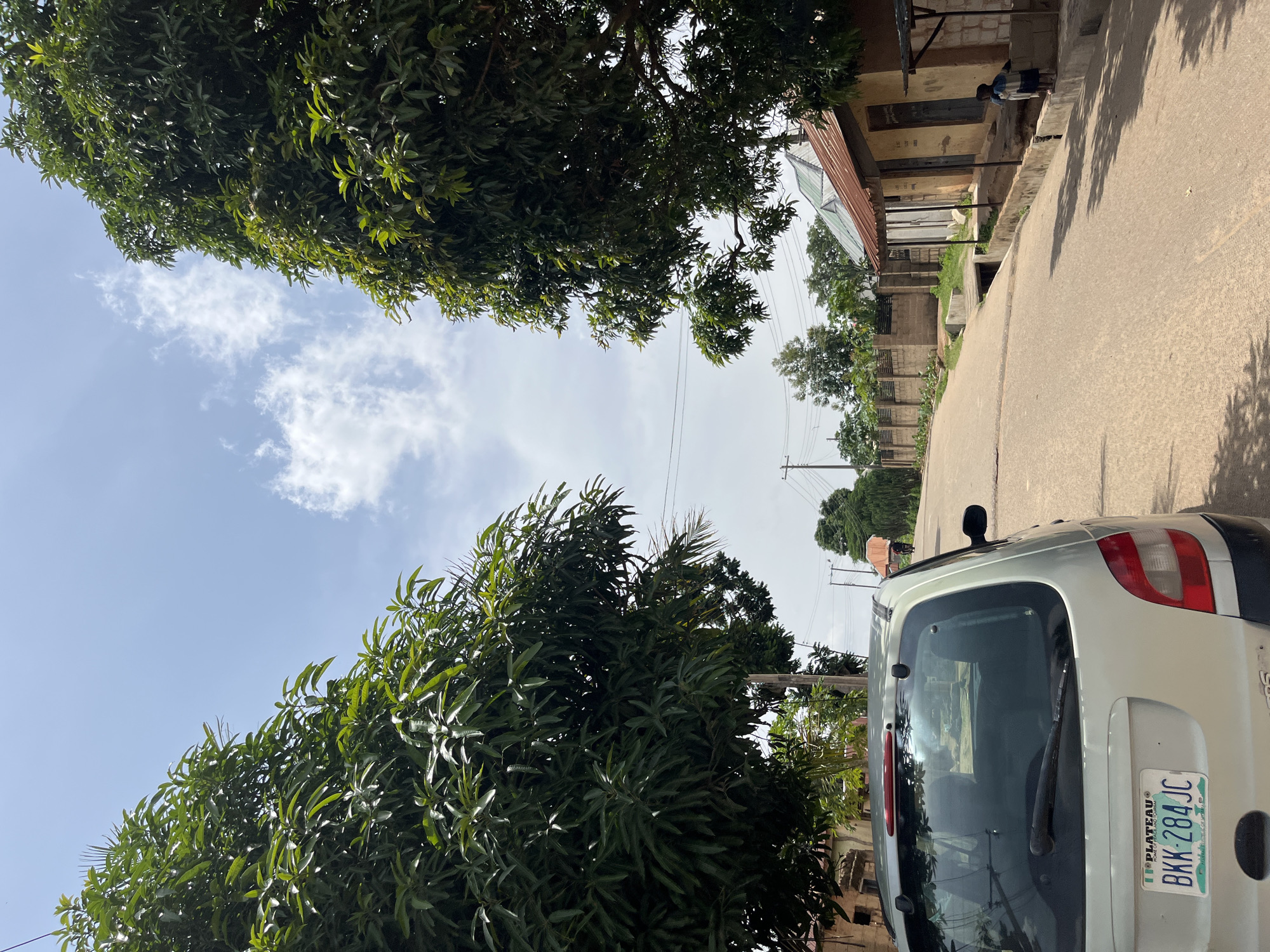 In Bukuru, Nigeria, a light-colored minivan is parked on the left side of a paved road. The minivan has a license plate reading "PLATEAU BKK-284 JC". The road curves gently uphill and to the right. On the right side of the road, there is a single-story building with a corrugated metal roof. Two young children, one wearing a blue and white outfit, are standing near the building. A person carrying a large object on their head, possibly a market stall or goods, is walking up the road in the distance. The scene is flanked by large, leafy trees, with power lines visible overhead. The sky is partly cloudy.