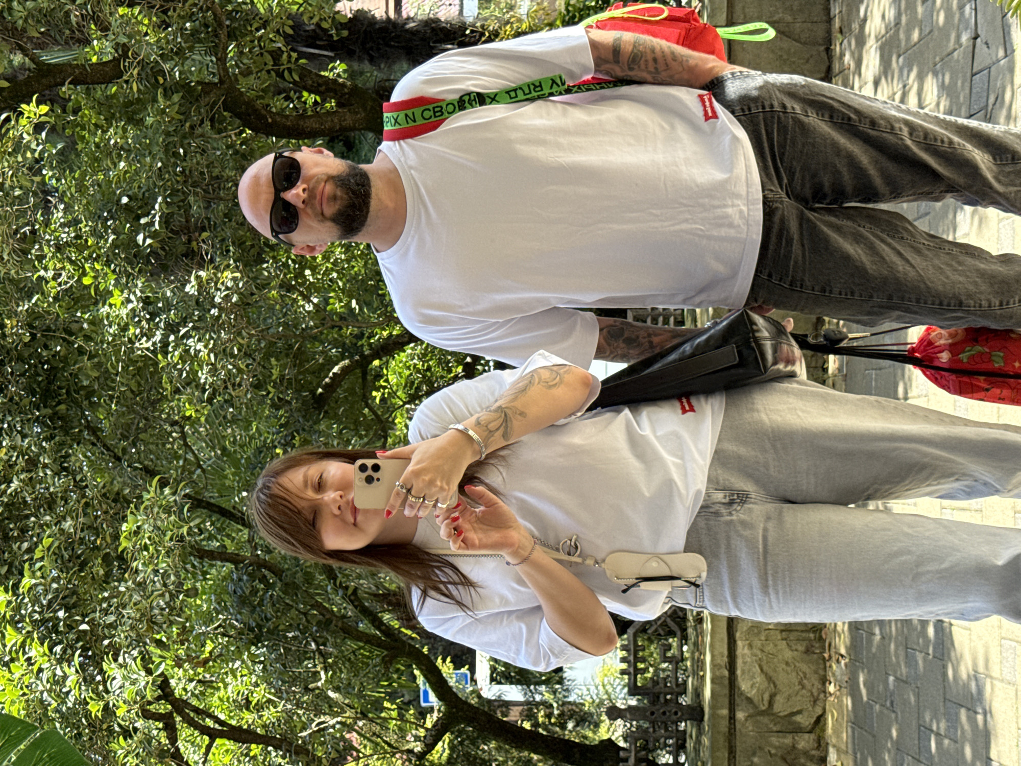 A middle-aged couple is taking a selfie in an outdoor urban environment with lush green trees in the background. The woman, on the left, is holding a smartphone and taking a picture of herself and the man. She is wearing a white t-shirt and light-wash jeans, and several rings on her fingers. The man, on the right, is wearing sunglasses, a white t-shirt, and dark-wash jeans. He has a beard and is carrying a red and green strap bag over his shoulder. A red bag with a floral pattern is hanging from his left hand. The ground is paved with rectangular stones, and a stone wall with an ornate metal fence is visible behind them. The scene is illuminated by bright sunlight, casting shadows on the ground. The environment suggests a park or a tree-lined street in a city. It is possible that the location is Sochi, Russia, given the presence of subtropical vegetation.